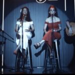 Cuarteto musical en directo con dos cantantes, guitarra acústica y percusión en escenario íntimo