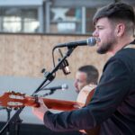 Cantante con guitarra acústica tocando en directo junto a otro músico al fondo