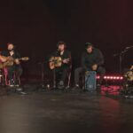 Grupo de rumba tocando en directo con guitarras, bajo, teclado y percusión en escenario oscuro
