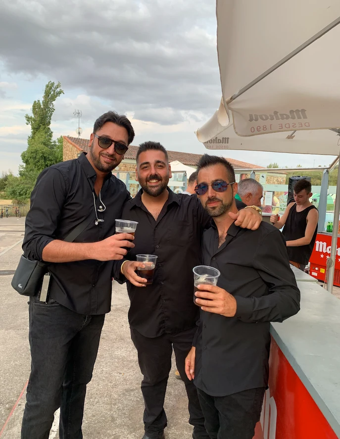 Tres hombres con camisas negras brindan con vasos en una barra al aire libre