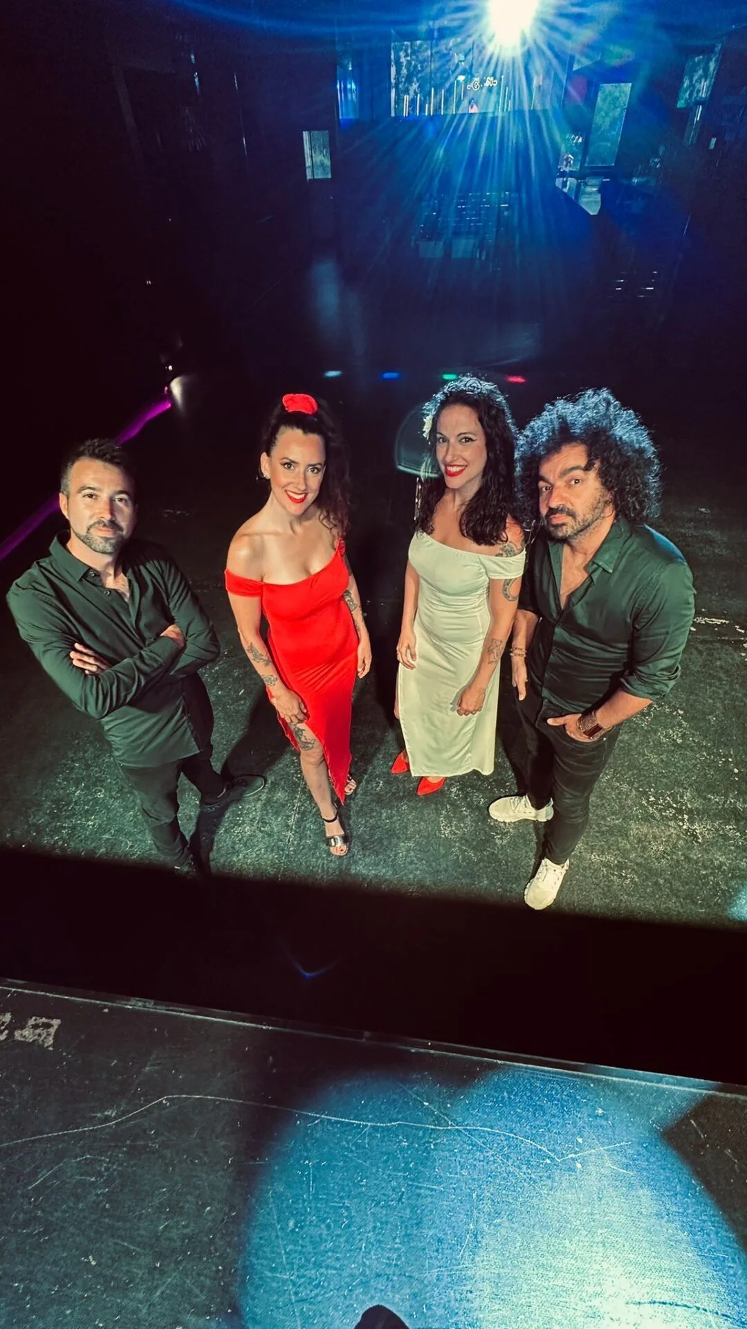 Cuatro músicos posan sobre un escenario con luces azules al fondo; dos mujeres con vestidos rojo y claro en el centro y dos hombres vestidos de oscuro a los lados, en un ambiente de sala de conciertos, ideal para rumba fusión y pop flamenco para eventos de empresa en Barcelona.
