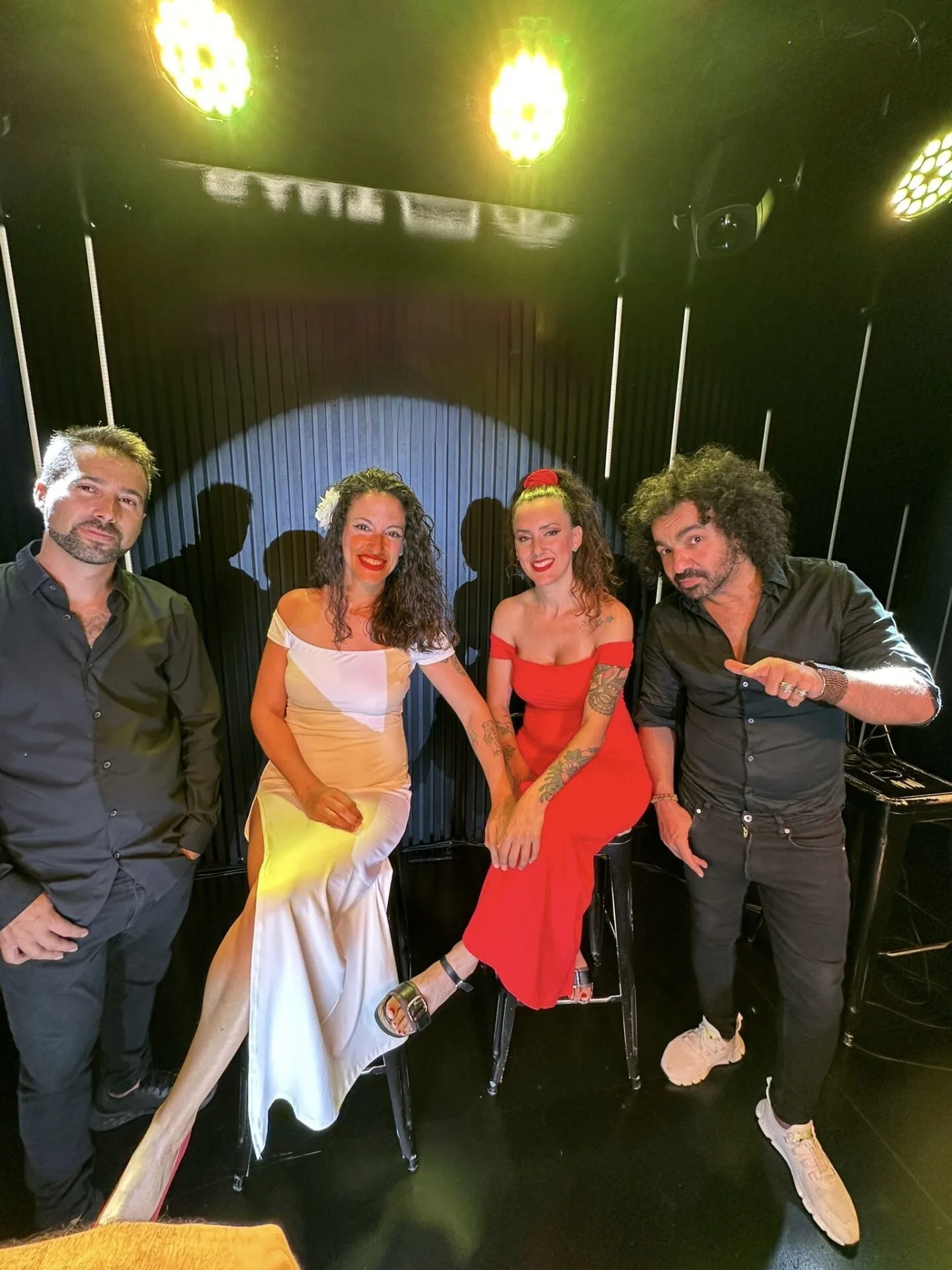 Cuatro integrantes posan en un escenario con fondo negro y focos amarillos encima; dos mujeres sentadas en taburetes (una con vestido blanco y otra con vestido rojo) y dos hombres de pie a los lados, una imagen pensada para promocionar rumba fusión y pop flamenco para eventos de empresa en Barcelona.