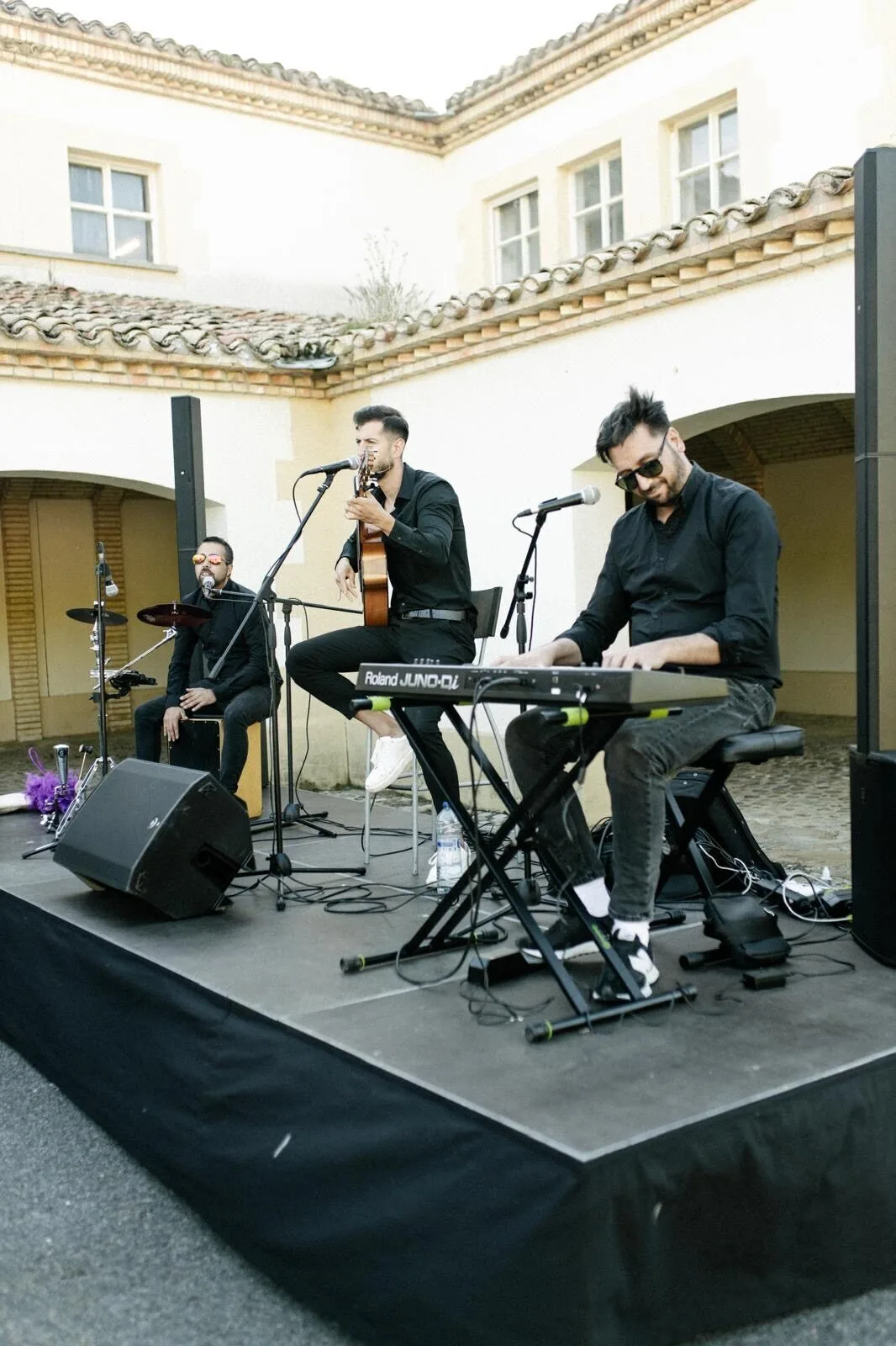 Tres músicos vestidos de negro actúan sobre un pequeño escenario exterior: a la derecha un teclista sentado toca un teclado, en el centro un cantante con guitarra acústica canta frente a un micrófono y a la izquierda un percusionista toca un cajón con una batería cerca; una escena ideal de flamenquito y pop flamenco en Barcelona para cumpleaños con buen rollo.