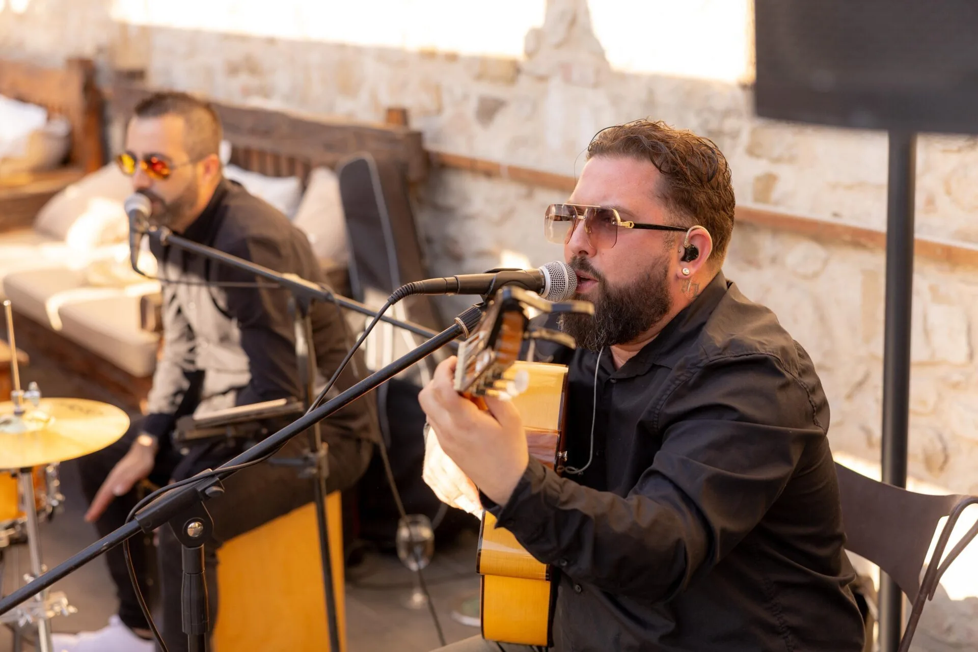 Primer plano de un cantante con guitarra española cantando en un micrófono, vestido de negro y con auriculares; al fondo se ve otro músico junto a una batería. La escena parece un set acústico para evento privado, con ambiente de noche en Barcelona y una rumba catalana con toque urbano.