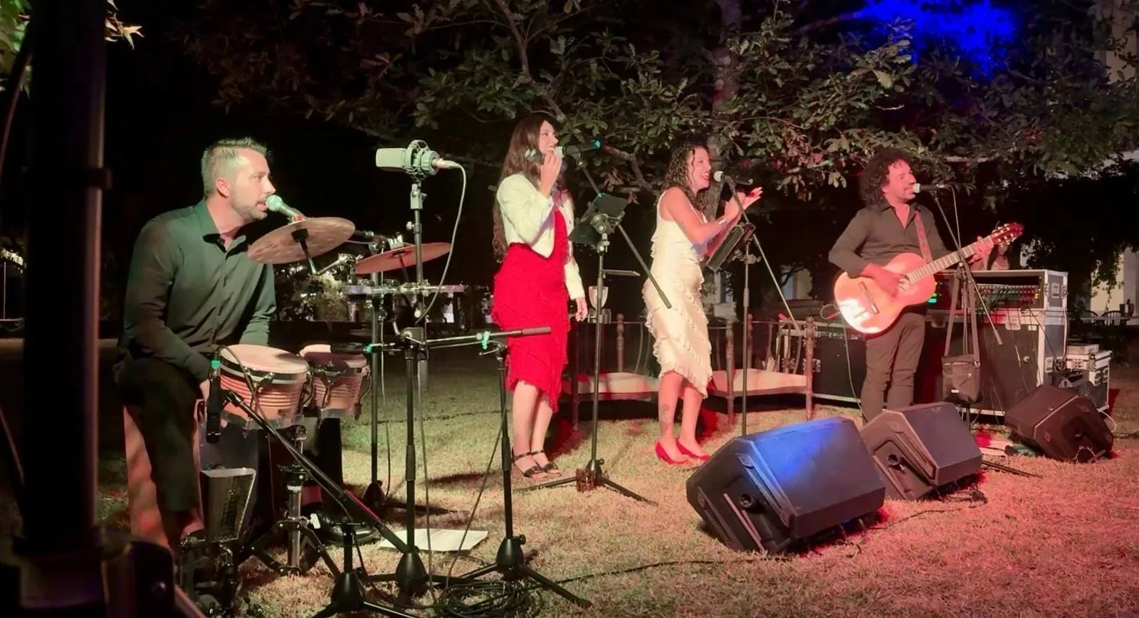 Escenario al aire libre de noche con cuatro músicos: un percusionista sentado tocando bongos, dos cantantes con micrófono en el centro y un guitarrista cantando a la derecha; delante hay monitores de sonido y cables sobre el césped, en un ambiente de rumba fusión y pop flamenco para eventos de empresa en Barcelona.
