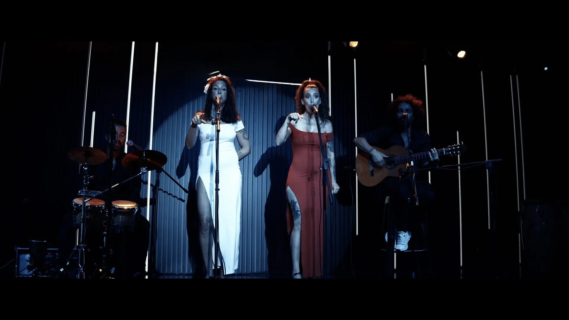 Dos cantantes con vestidos largos, uno blanco y otro rojo, cantan en dos micrófonos sobre un escenario con luces verticales; a la izquierda se ve un percusionista con congas y batería, y a la derecha un guitarrista con guitarra española, una imagen adecuada para rumba fusión y pop flamenco para eventos de empresa en Barcelona.