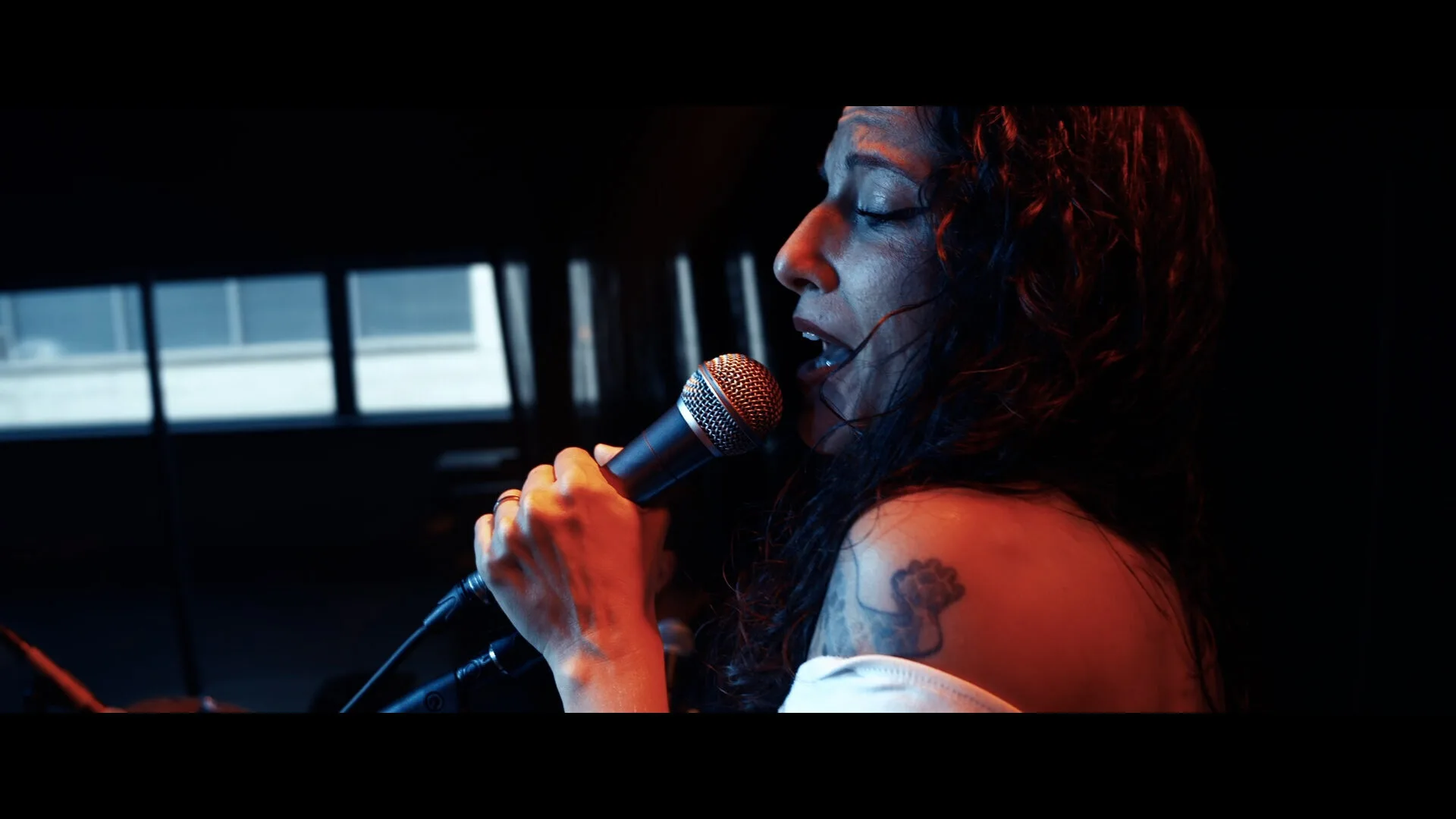 Plano lateral de una cantante con el pelo oscuro y rizado, sosteniendo un micrófono muy cerca de la boca mientras canta; la luz roja ilumina su rostro y hombro, donde se aprecia un tatuaje. Fondo interior desenfocado con ventanas, ideal para ilustrar rumba fusión y pop flamenco para eventos de empresa en Barcelona.