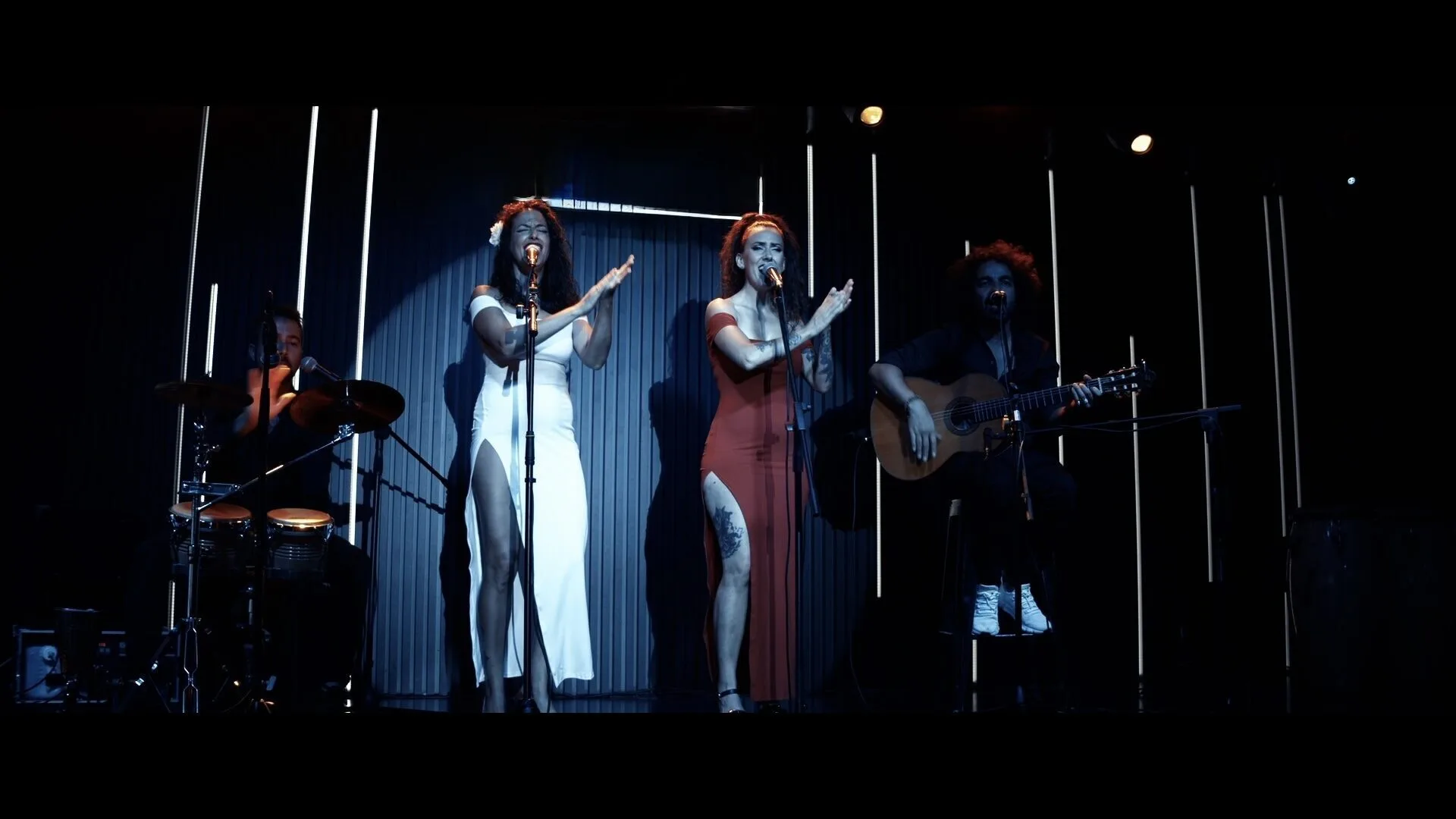 Dos cantantes en el centro del escenario aplauden y cantan frente a sus micrófonos; una lleva un vestido blanco y la otra un vestido rojo con abertura. A la izquierda se ve un percusionista con batería y, a la derecha, un guitarrista sentado con guitarra española. Iluminación oscura con barras de luz verticales, en un montaje típico de rumba fusión y pop flamenco para eventos de empresa en Barcelona.