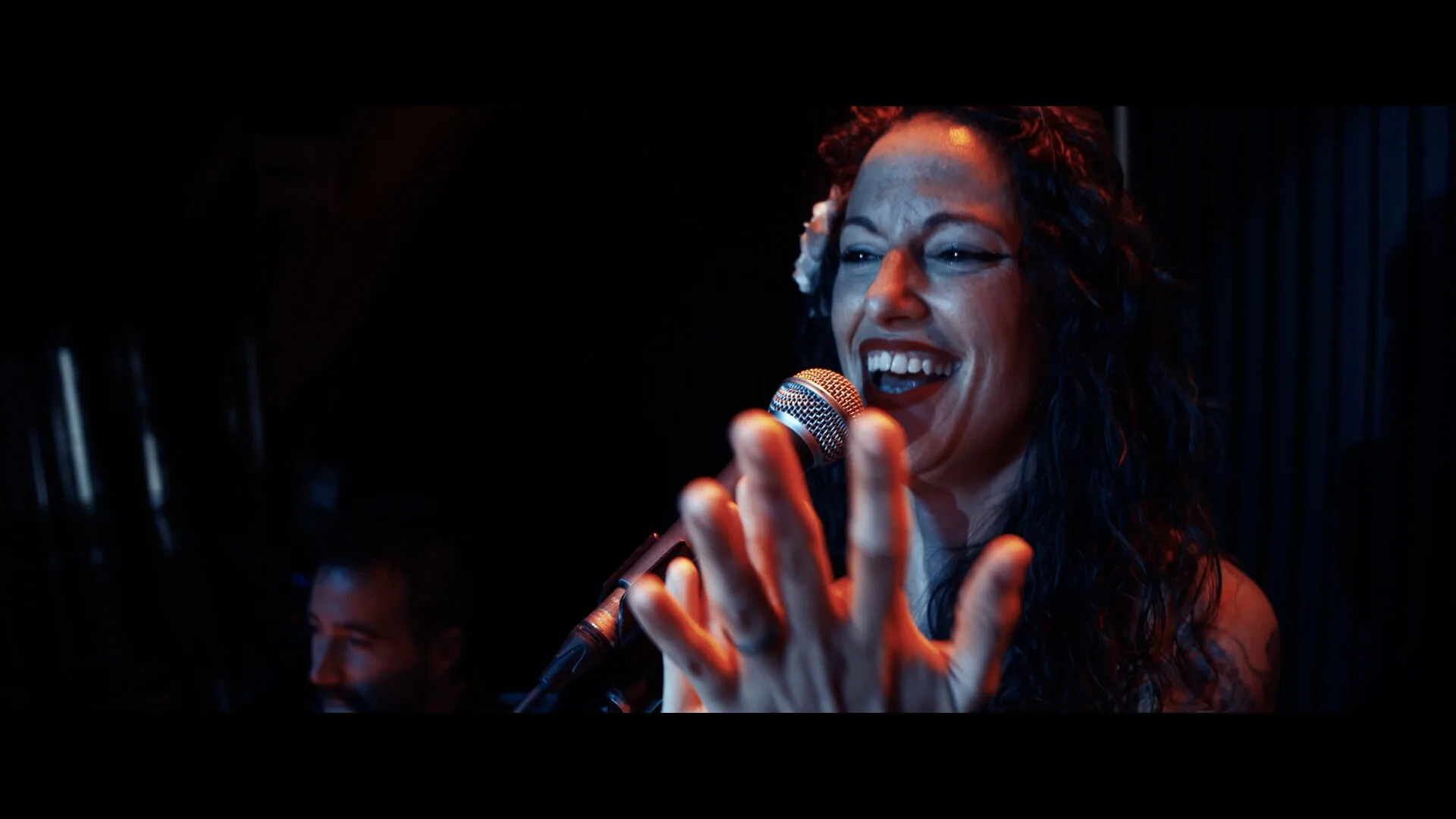 Primer plano de una cantante sonriendo mientras canta a un micrófono y marca el ritmo con las manos; iluminación cálida en un escenario oscuro, ideal para rumba fusión y pop flamenco en eventos de empresa en Barcelona.