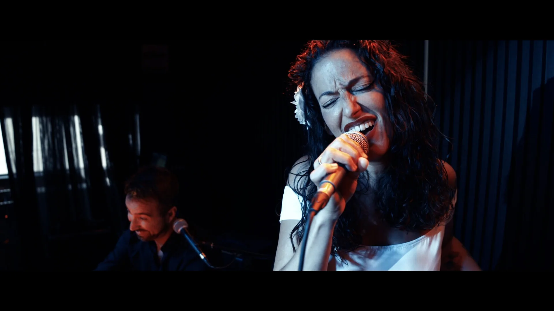 Primer plano de una cantante con el pelo rizado y una flor blanca en el pelo, cantando con los ojos cerrados y sujetando un micrófono; al fondo, desenfocado, aparece otro músico con micrófono en un ambiente interior con luces bajas, una imagen adecuada para promocionar rumba fusión y pop flamenco para eventos de empresa en Barcelona.