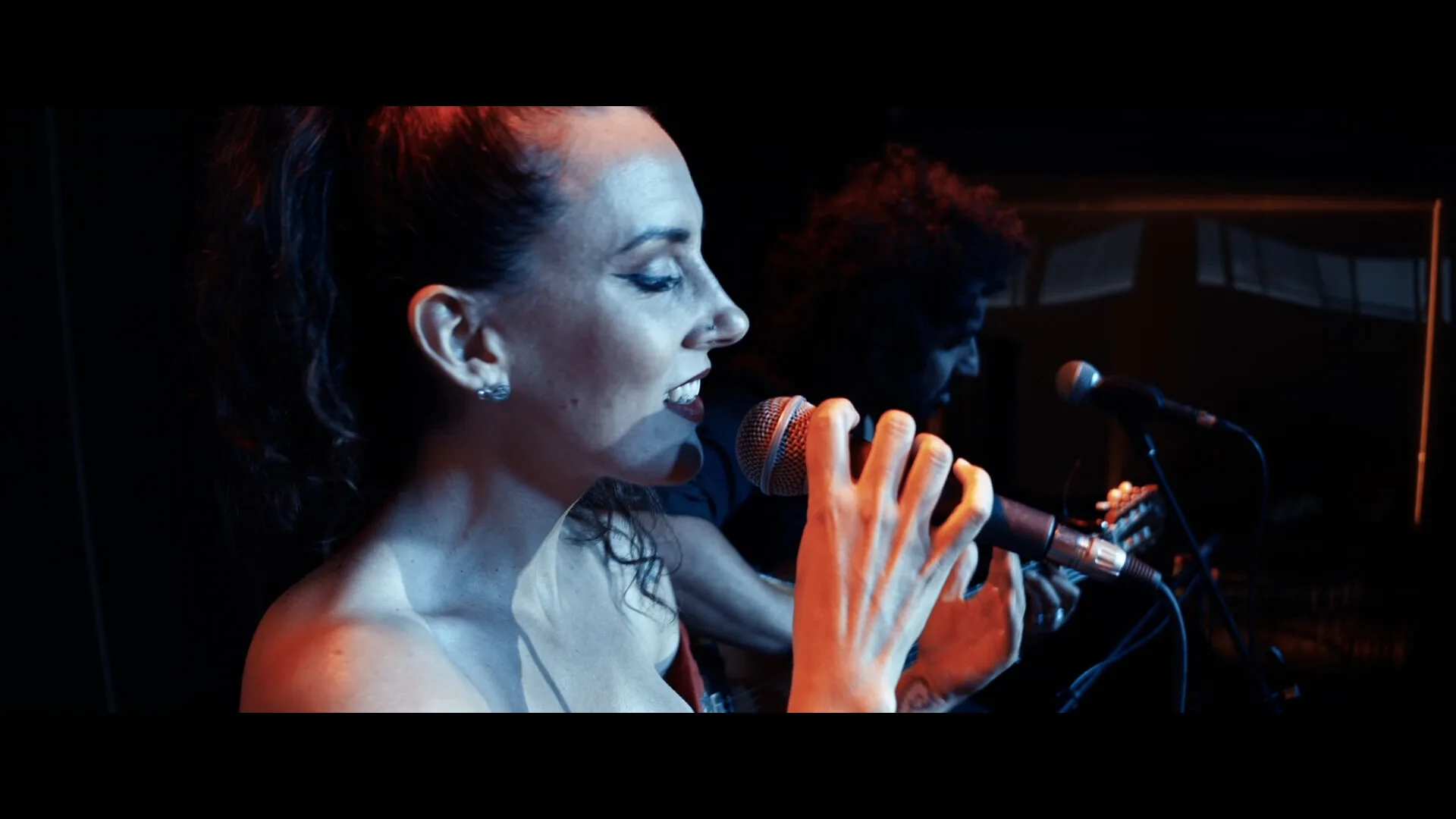 Primer plano de una cantante de perfil sujetando un micrófono y cantando en un escenario con luz azul y cálida; al fondo se intuye otro músico y una guitarra. Imagen asociada a rumba fusión y pop flamenco para eventos de empresa en Barcelona.