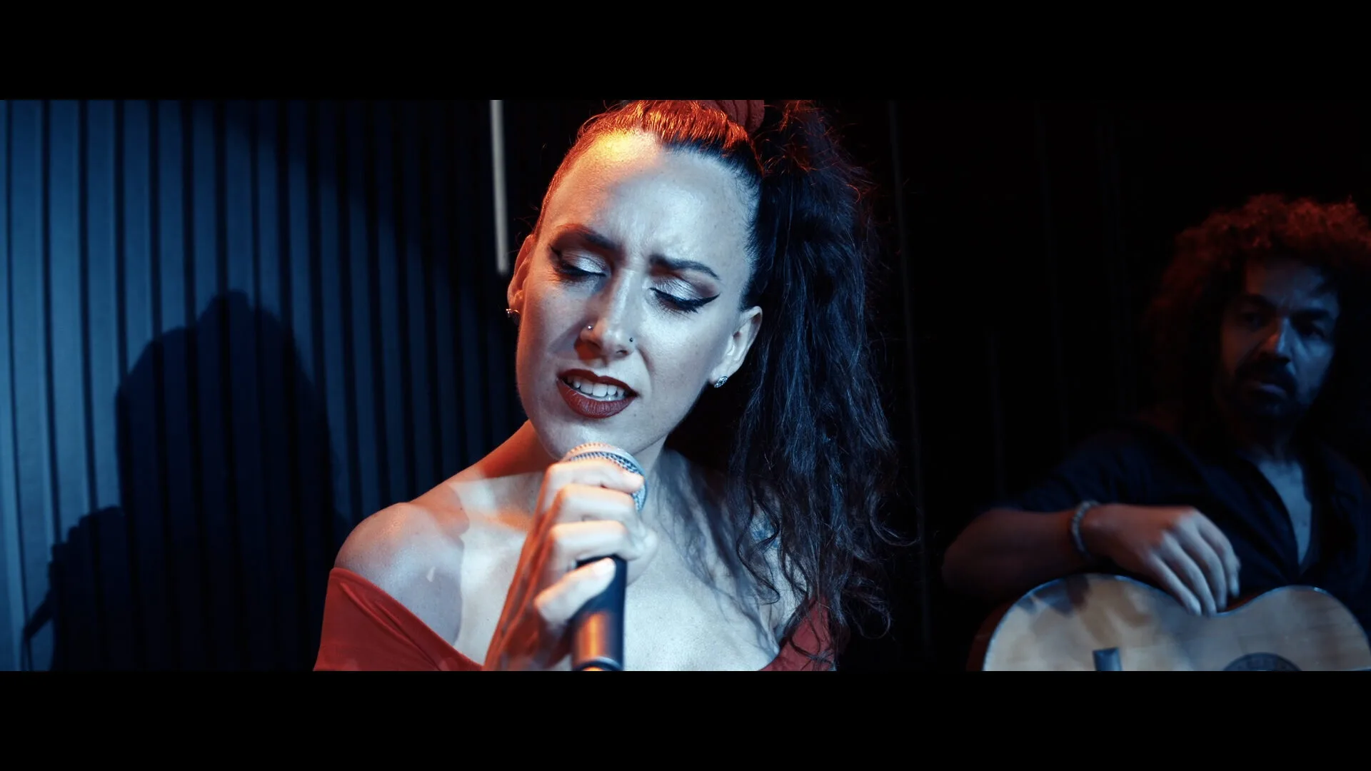 Primer plano de una cantante con coleta alta y maquillaje marcado, sujetando un micrófono mientras canta con los ojos cerrados; al fondo, desenfocado, aparece un músico con guitarra. Iluminación azul y cálida en un escenario interior, ideal para rumba fusión y pop flamenco para eventos de empresa en Barcelona.