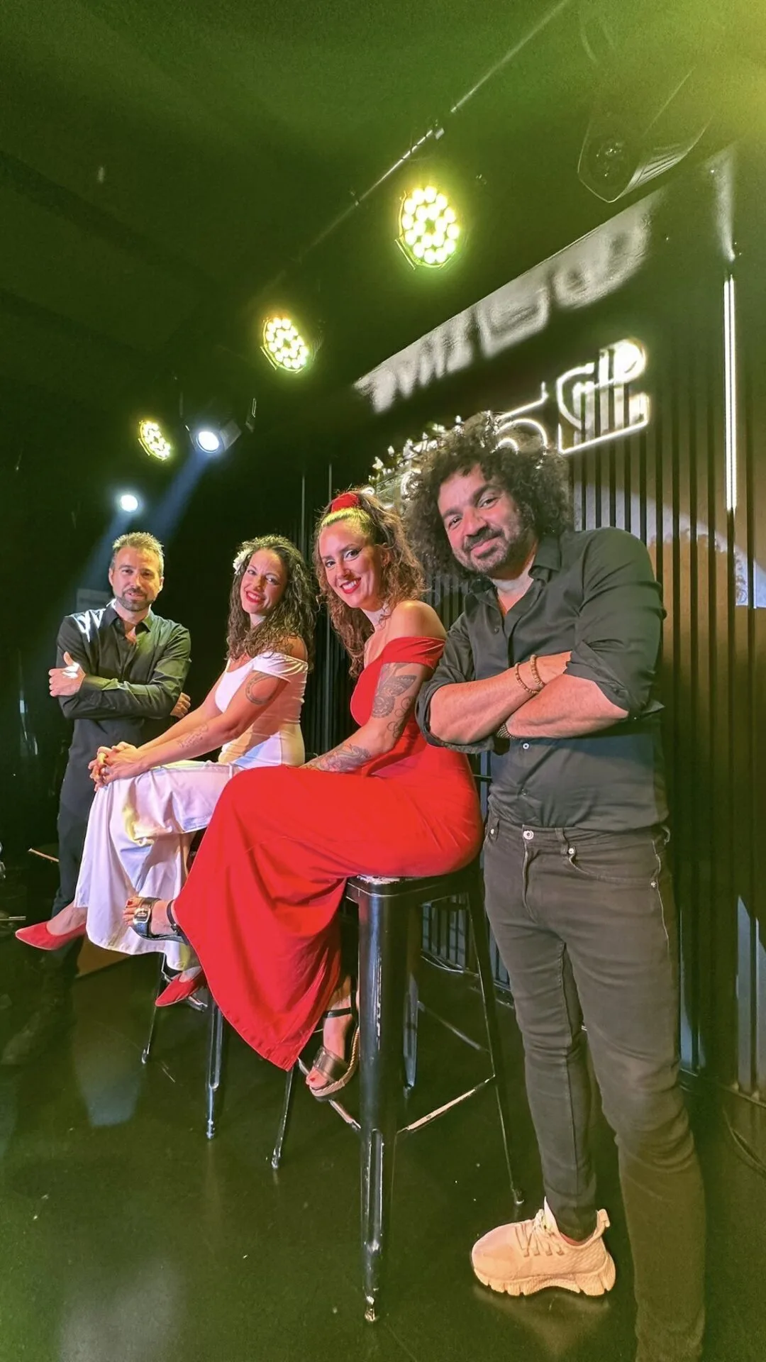Cuatro músicos posan en un escenario interior con focos amarillos arriba; dos mujeres sentadas en taburetes (una con vestido blanco y otra con vestido rojo) y dos hombres de pie a los lados, todos mirando a cámara. Imagen promocional de rumba fusión y pop flamenco para eventos de empresa en Barcelona.