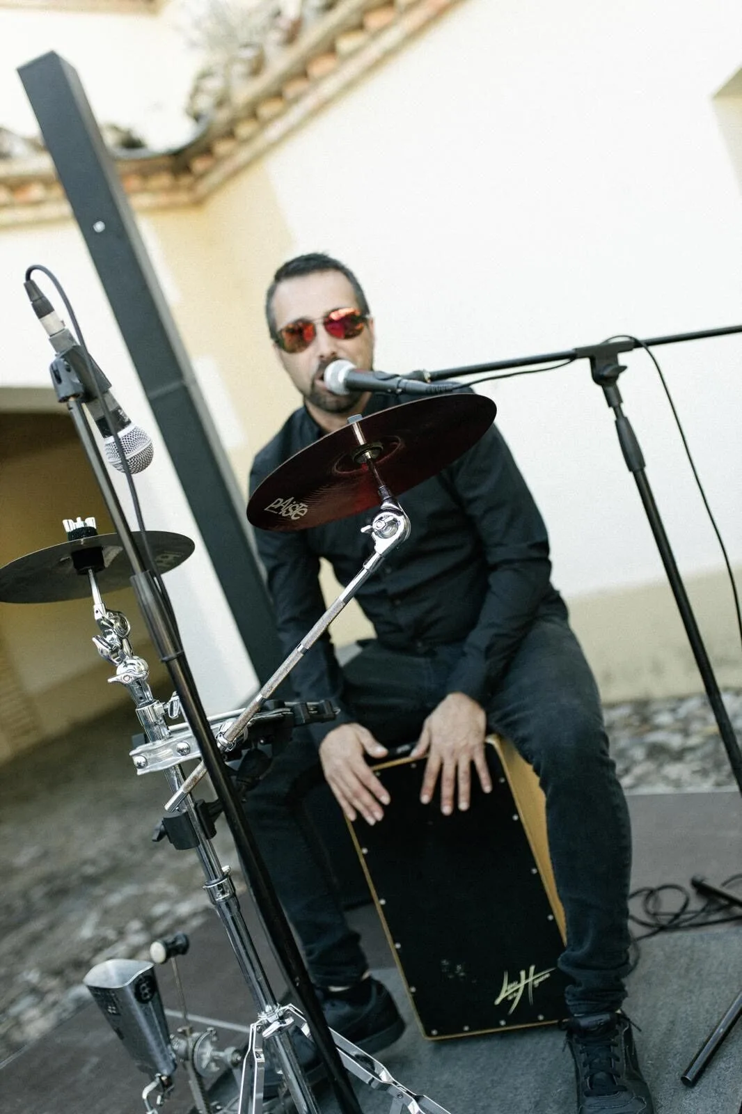 Músico sentado tocando un cajón y cantando frente a un micrófono, con un plato de batería y soportes en primer plano, en un montaje exterior; imagen ideal para flamenquito y pop flamenco en Barcelona para cumpleaños con buen rollo.