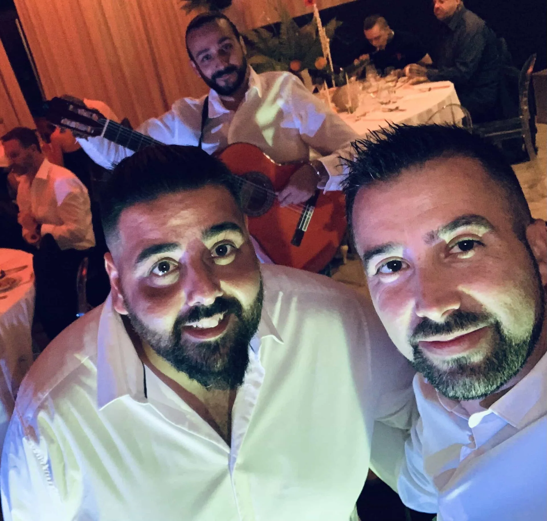 Selfie de dos hombres con camisa blanca y barba sonrientes en un salón de celebración; al fondo se ve un guitarrista con guitarra española junto a mesas vestidas, una escena de rumba que arranca sonrisas en tu boda en Tarragona.