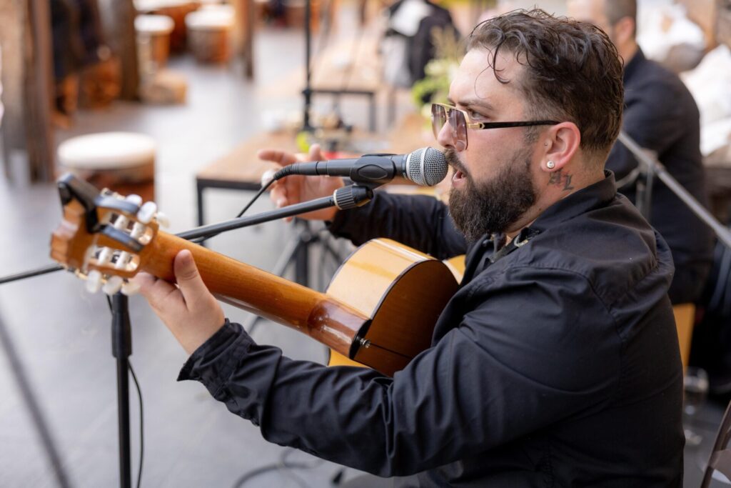 Músico com óculos escuros canta ao microfone enquanto toca violão espanhol, visto de perfil num espaço de evento com mesas ao fundo, num ambiente que sugere rumba catalã com toque urbano para a noite em Barcelona.
