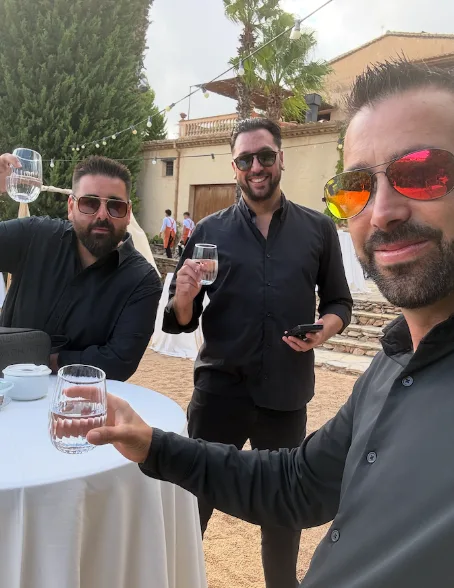 Tres hombres vestidos de negro brindan con vasos de agua junto a una mesa alta con mantel blanco, en un patio exterior con guirnaldas de luces y una palmera; una escena de celebración que encaja con una rumba que arranca sonrisas en tu boda en Tarragona.