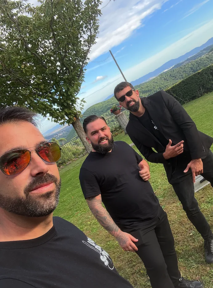 Tres hombres posan para una foto al aire libre en un jardín con césped y vistas a montañas; uno aparece en primer plano con gafas de sol y barba, y los otros dos están detrás, vestidos de negro. Imagen asociada a una rumba catalana con toque urbano para la noche en Barcelona, ideal para bodas y eventos privados.