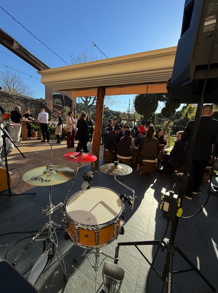 En primer plano se ve una batería montada sobre un escenario, con platos y caja, y varios pies de micro y cables alrededor. Al fondo, en una terraza exterior con pérgola y cielo despejado, hay grupos de invitados sentados y de pie conversando, como en una celebración. La escena sugiere un montaje musical para un evento, ideal para una rumba catalana con toque urbano para la noche en Barcelona.