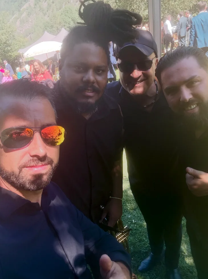 Selfie de cuatro hombres mirando a cámara en un evento al aire libre, con gente y carpas al fondo; dos llevan gafas de sol y uno gorra. La escena sugiere un ambiente de celebración, ideal para una rumba catalana con toque urbano para la noche en Barcelona.