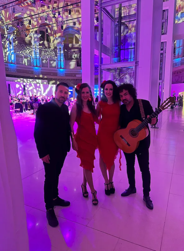 Cuatro músicos posan en un gran salón de eventos iluminado en tonos morados y azules; dos mujeres con vestidos rojos y dos hombres de negro, uno de ellos sosteniendo una guitarra española. Al fondo se ven mesas con invitados y una arquitectura interior amplia, una escena asociada a rumba fusión y pop flamenco para eventos de empresa en Barcelona.