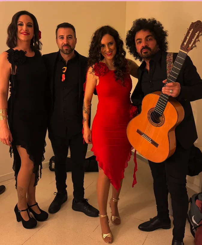 Cuatro integrantes de un grupo musical posan en interior: dos mujeres (una con vestido negro y otra con vestido rojo) y dos hombres vestidos de negro; el hombre de la derecha sostiene una guitarra española. Imagen tipo backstage o camerino, adecuada para promocionar rumba fusión y pop flamenco para eventos de empresa en Barcelona.