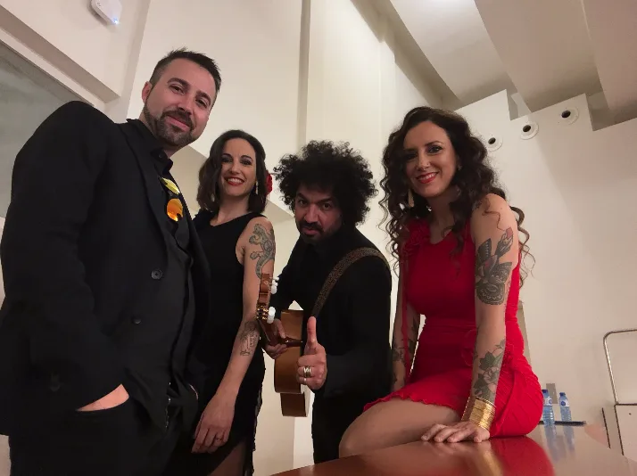 Foto de cuatro integrantes de un grupo musical posando en un interior blanco: un hombre con traje negro, dos mujeres con vestidos (una rojo y otra negro) y un guitarrista con el instrumento en la mano; imagen tipo backstage o posado, relacionada con rumba fusión y pop flamenco para eventos de empresa en Barcelona.