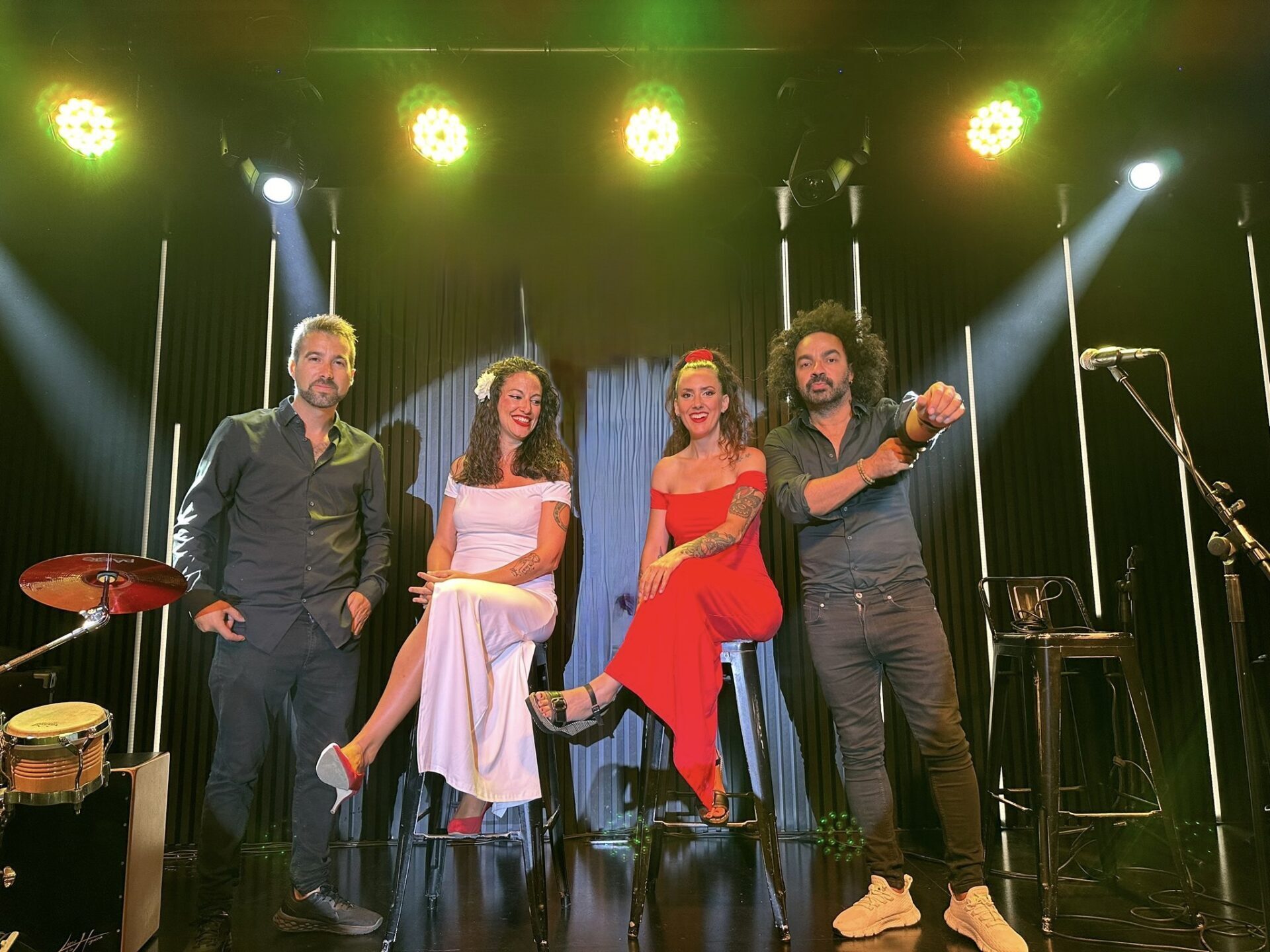 Quatro músicos posam num palco com cortinas pretas e focos amarelos e brancos no teto, dois sentados em bancos altos e dois de pé; à esquerda vê se parte de uma bateria e, à direita, um microfone num tripé, num ambiente típico de rumba fusão e pop flamenco para eventos de empresa em Barcelona.