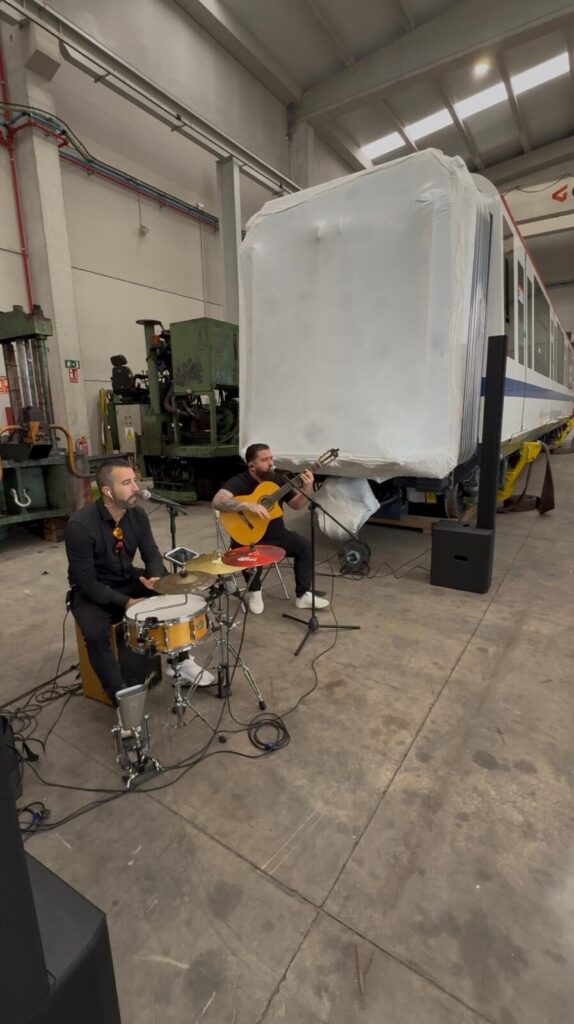 Dois músicos num espaço industrial, um a cantar ao microfone enquanto toca percussão com tarola e prato, e outro sentado a tocar guitarra espanhola com microfone; ao fundo vê se uma carruagem de comboio coberta por plástico, num ambiente de rumba catalã com toque urbano para a noite em Barcelona.