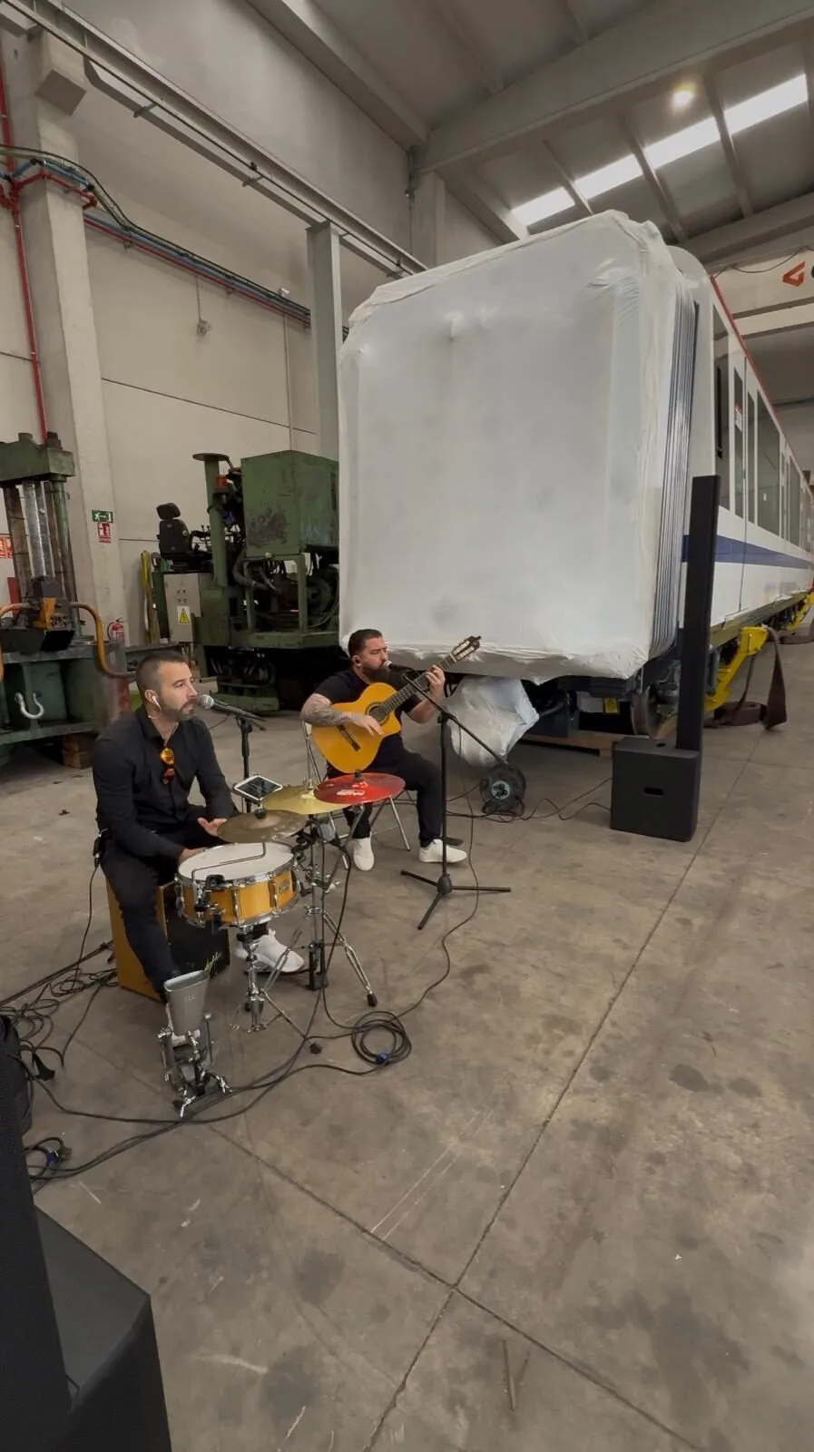 Dos músicos actúan en un espacio industrial: a la izquierda, un percusionista sentado toca una caja con plato y canta a un micrófono; a la derecha, un guitarrista también canta. Hay altavoces y cables en el suelo, y detrás se ve un gran vagón o estructura blanca dentro de un taller. La escena transmite una rumba catalana con toque urbano para la noche en Barcelona.