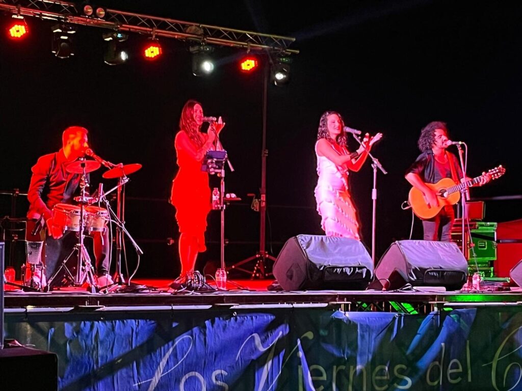 Banda no palco com iluminação vermelha, com um percussionista à esquerda, duas cantoras ao centro com microfones e um guitarrista e cantor à direita, num cenário de rumba fusão e pop flamenco para eventos de empresa em Barcelona.