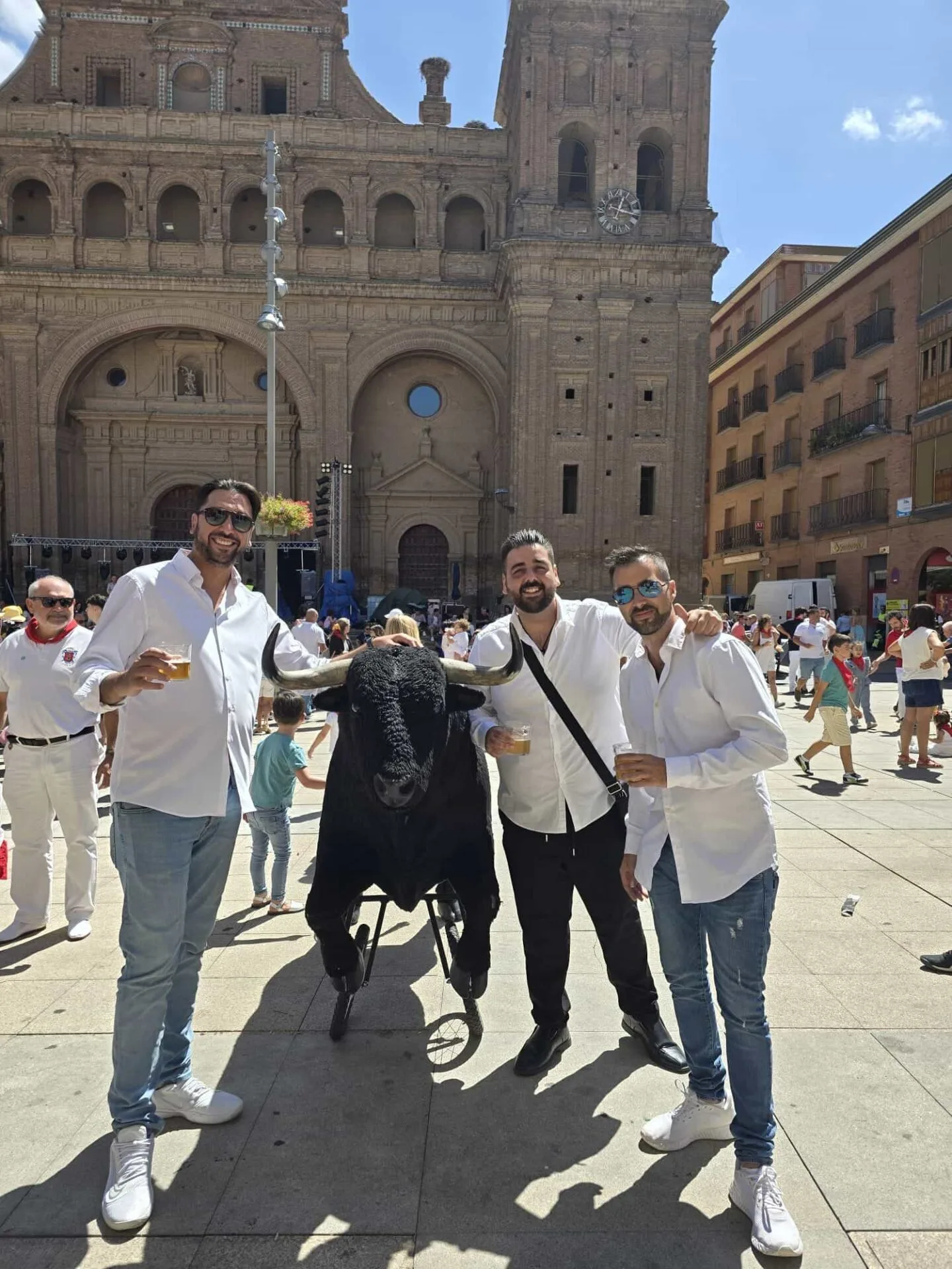 Tres hombres con camisas blancas posan sonrientes en una plaza concurrida, junto a una figura de toro negro con cuernos; al fondo se ve una gran fachada de iglesia con torre y reloj. La escena transmite un ambiente festivo, ideal para una rumba que arranca sonrisas en tu boda en Tarragona.