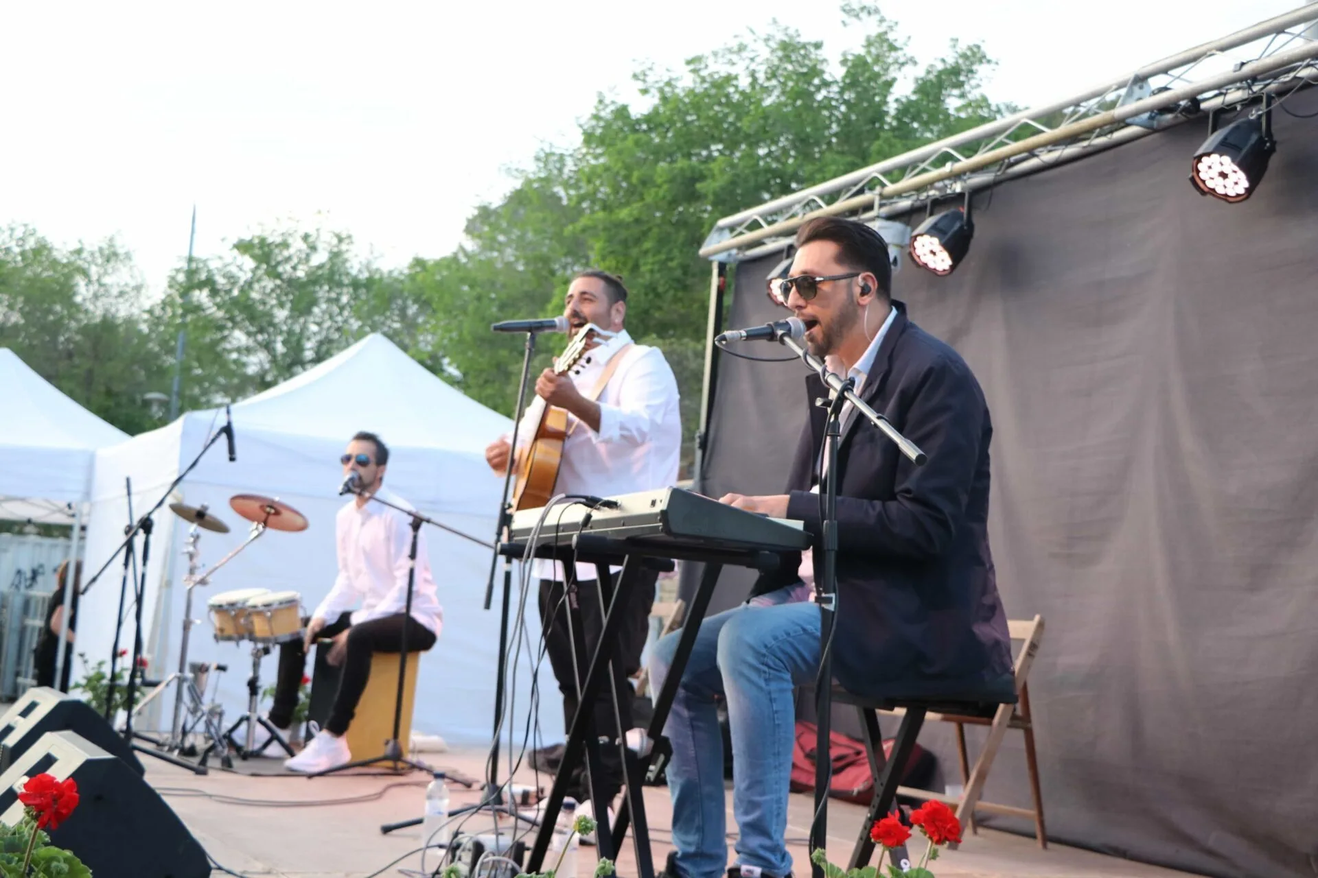 Tres músicos tocan sobre un escenario al aire libre: en primer plano, un cantante con gafas canta a un micrófono mientras toca un teclado; detrás, otro músico canta y toca una guitarra española; a la izquierda, un percusionista sentado acompaña con un cajón y platos. Hay focos y una lona negra de fondo, con árboles y carpas blancas alrededor, en un ambiente de rumba que arranca sonrisas en tu boda en Tarragona.