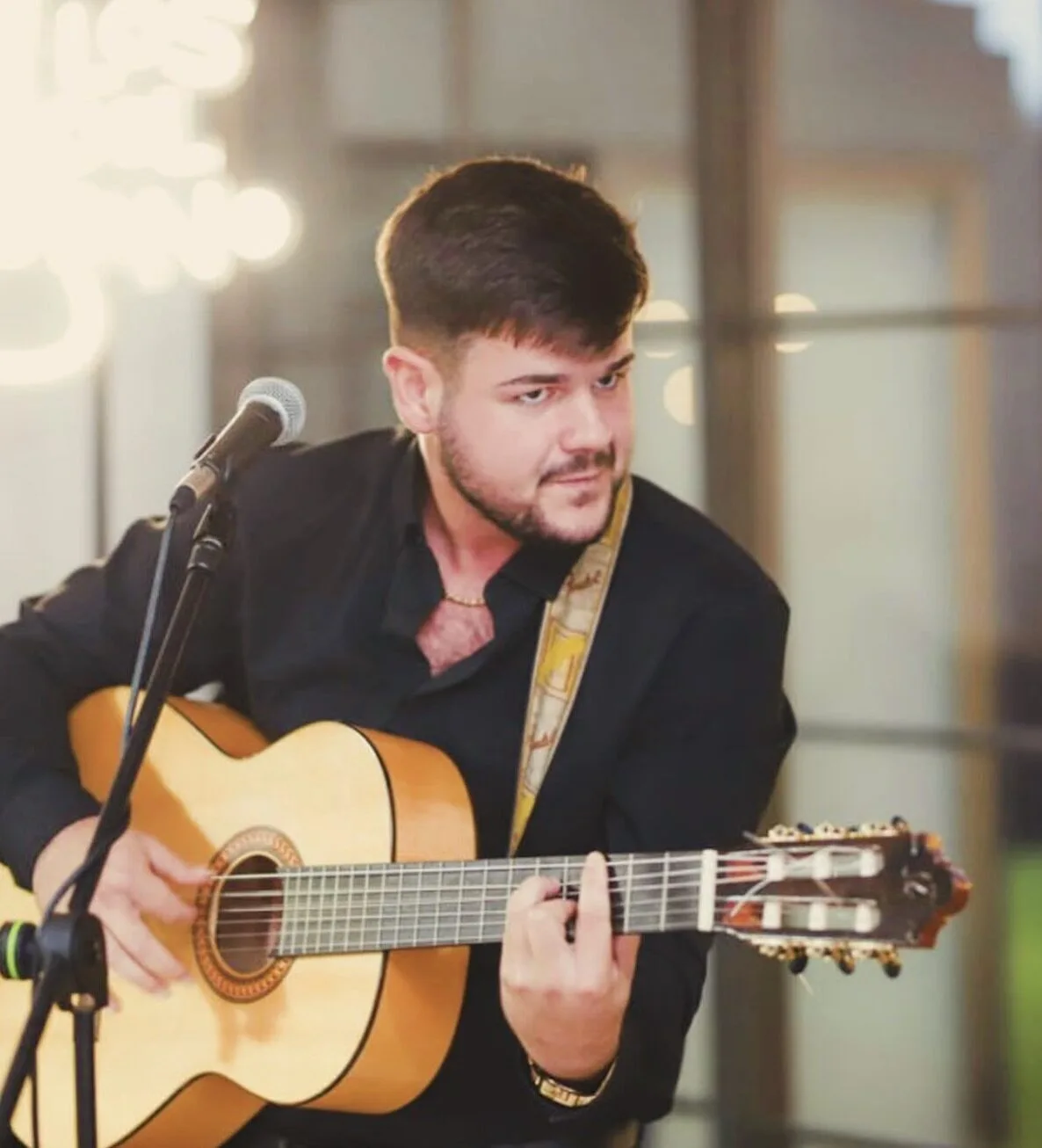 Primer plano de un guitarrista con camisa negra tocando una guitarra española junto a un micrófono, en un interior con luces desenfocadas; imagen ideal para rumba y flamenquito para una boda con fiesta en Barcelona.