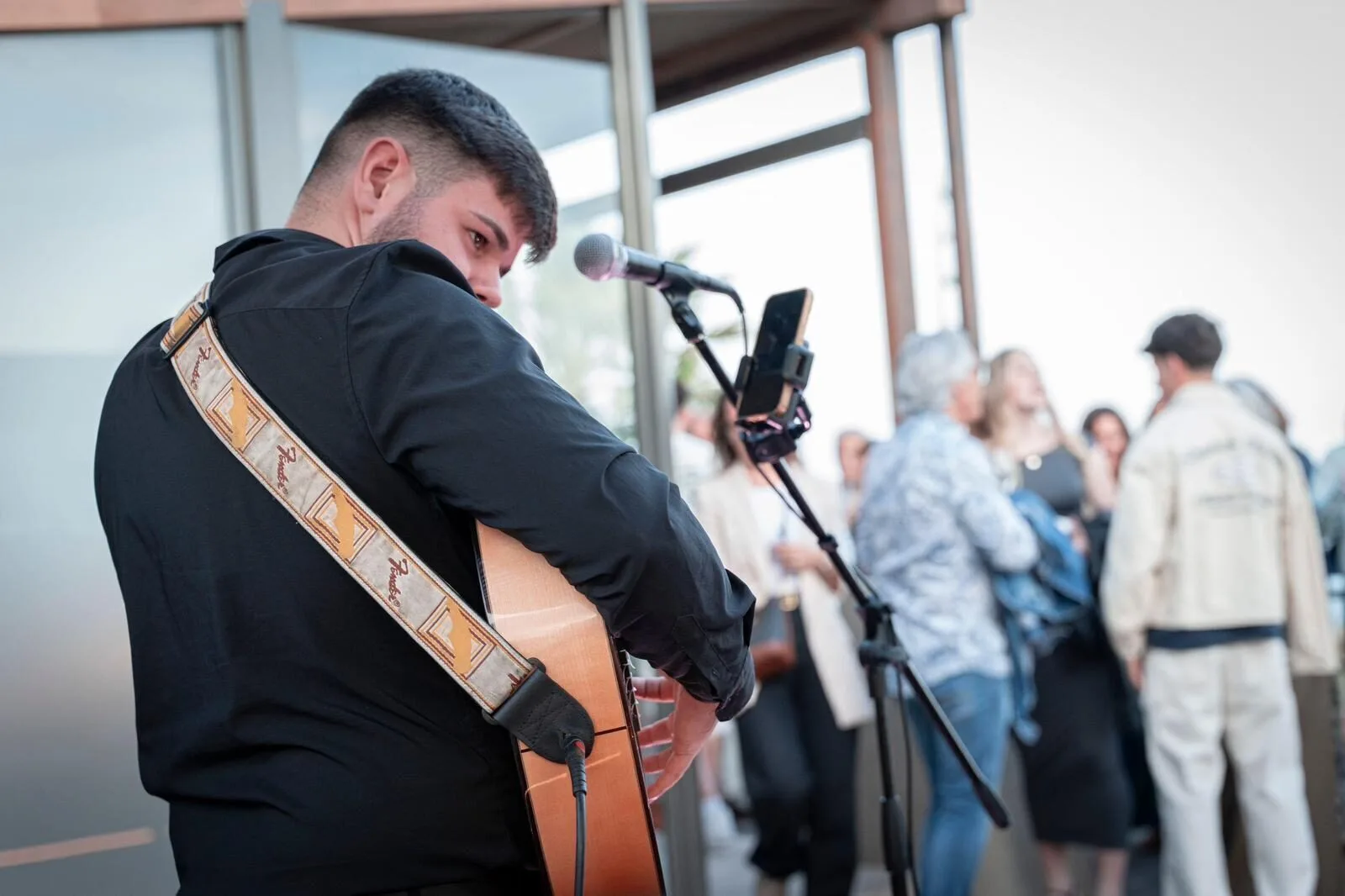 Primer plano de un guitarrista con camisa negra y la guitarra colgada al hombro, tocando junto a un micrófono en un espacio interior luminoso; al fondo se ven invitados charlando y moviéndose, ambiente de rumba y flamenquito para una boda con fiesta en Barcelona.