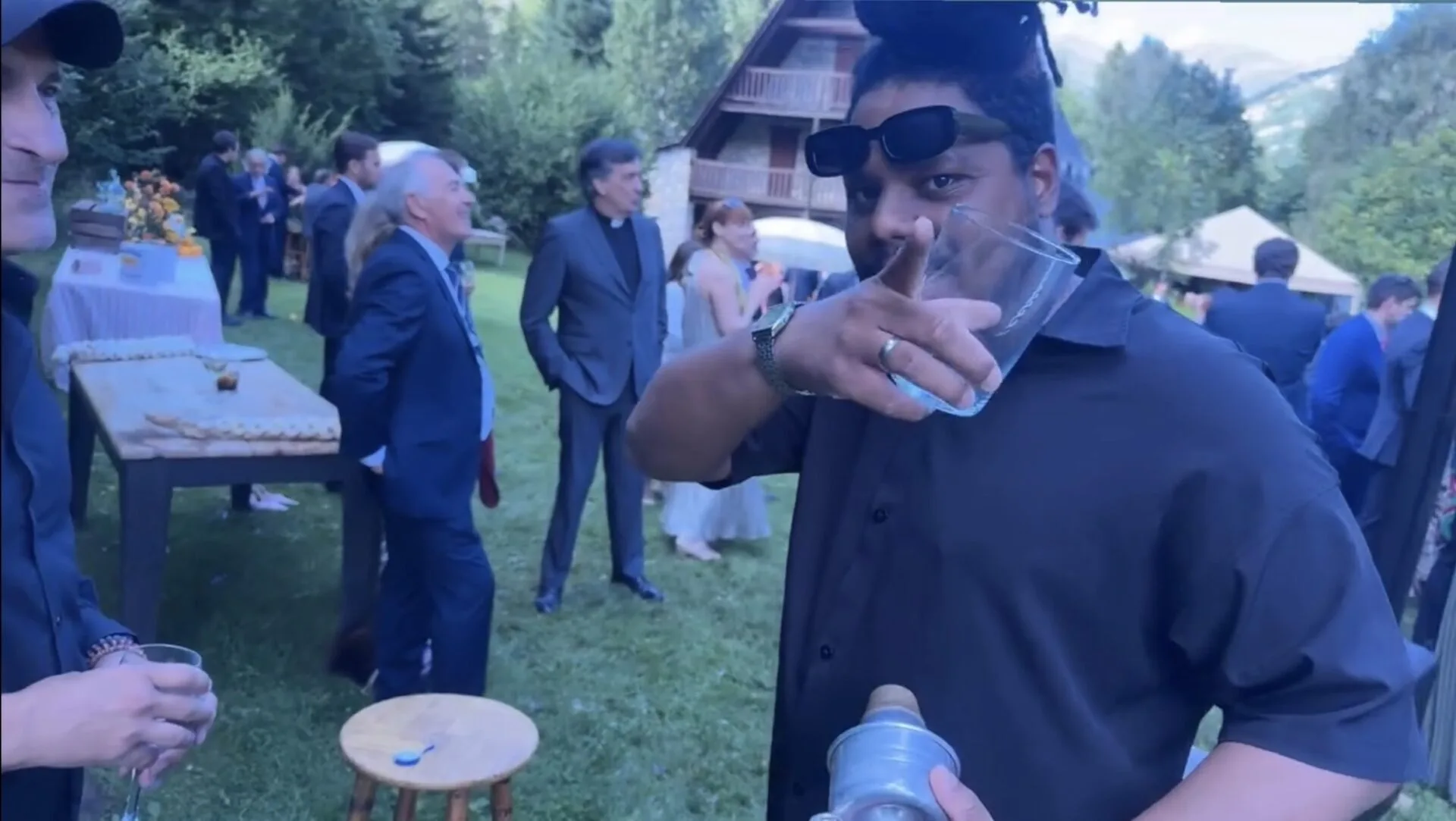En primer plano, un invitado con camisa oscura y gafas de sol en la cabeza sostiene un vaso de plástico y hace un gesto hacia la cámara durante un cóctel al aire libre; al fondo se ven más personas conversando junto a una mesa alta en un jardín con una casa de madera, un ambiente de celebración que encaja con una rumba catalana con toque urbano para la noche en Barcelona.