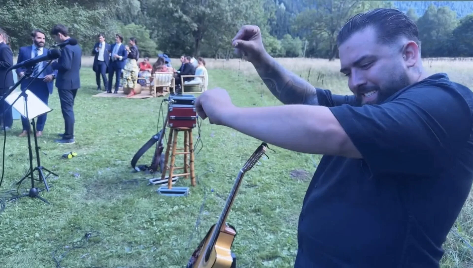 Primer plano de un músico con camiseta negra sonriendo y levantando el brazo, con una guitarra colgada; al fondo se ven micrófonos y varios invitados vestidos de traje y vestido conversando y sentados en un jardín, en un ambiente de celebración que encaja con una rumba catalana con toque urbano para la noche en Barcelona.
