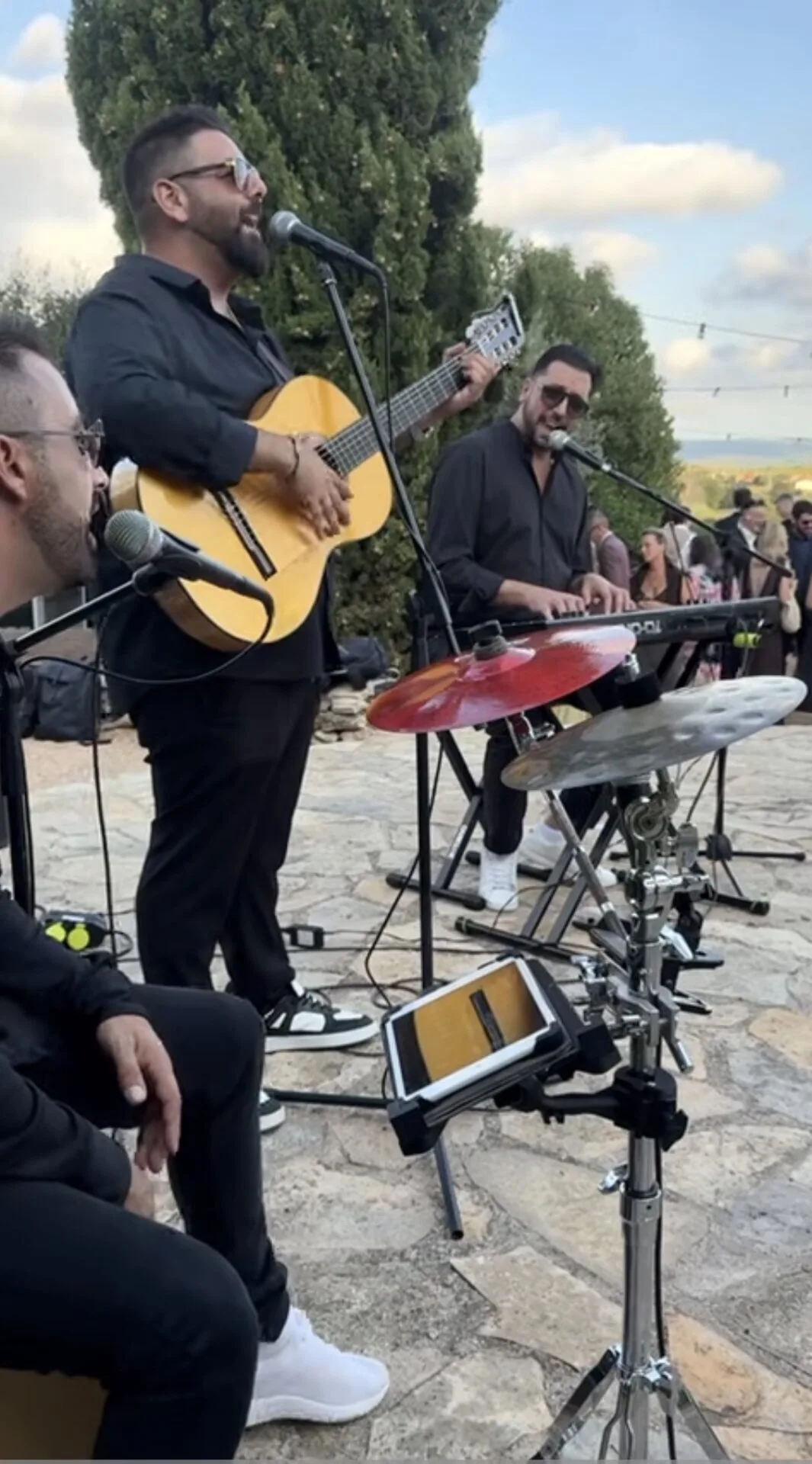 Grupo de rumba con tres músicos vestidos de negro tocando en una terraza al aire libre: un cantante con guitarra frente a un micrófono, un teclista cantando y parte de una batería con platos en primer plano; al fondo se ven invitados, en un ambiente de celebración, una rumba que arranca sonrisas en tu boda en Tarragona.