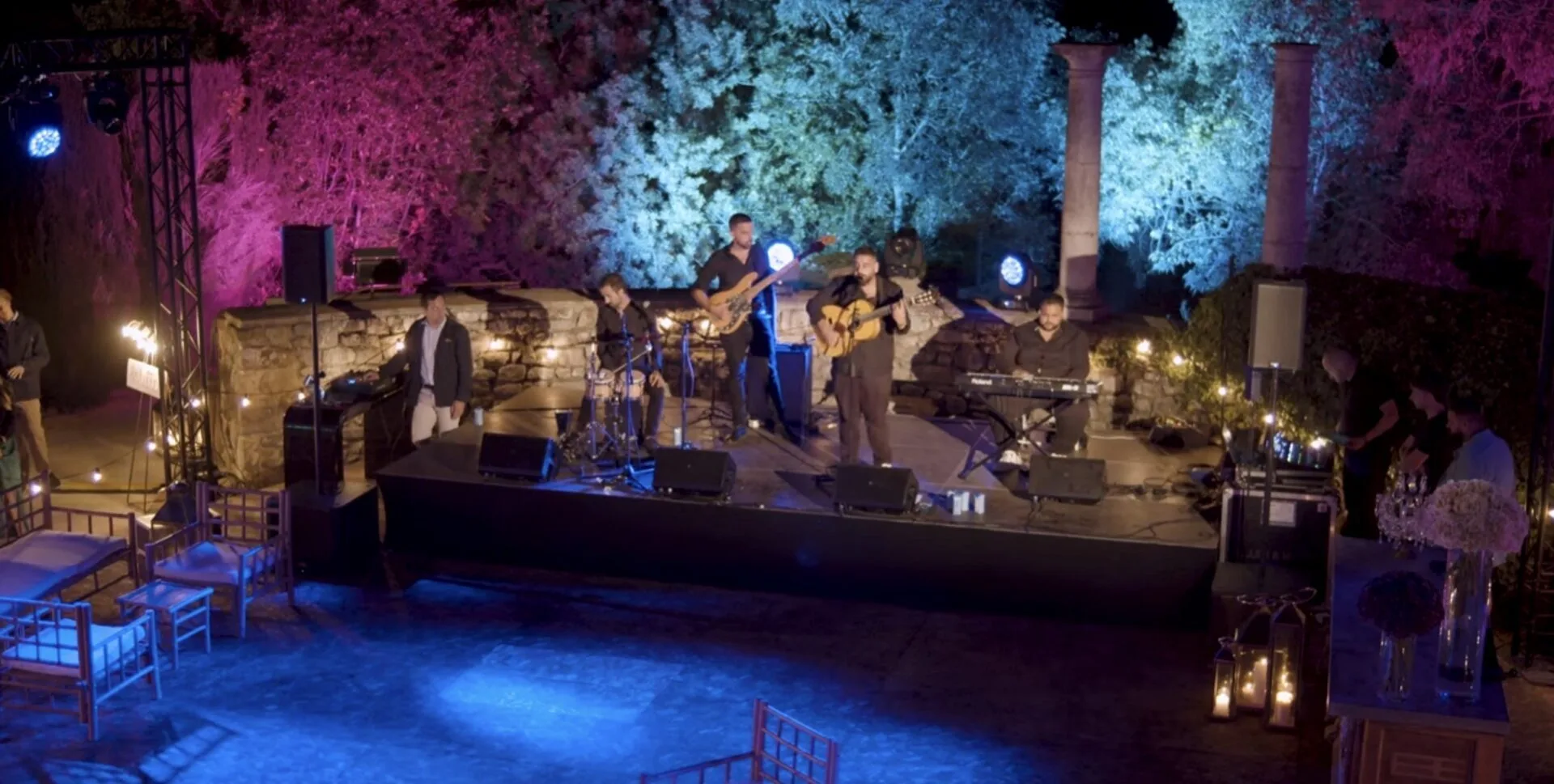Escenario exterior de noche en una finca, con un grupo de música española tocando: cantante con guitarra al frente, bajista detrás, batería y teclista a los lados, mientras el público observa desde la derecha. Las luces moradas y azules iluminan los árboles y crean un ambiente festivo, una rumba que arranca sonrisas en tu boda en Tarragona.