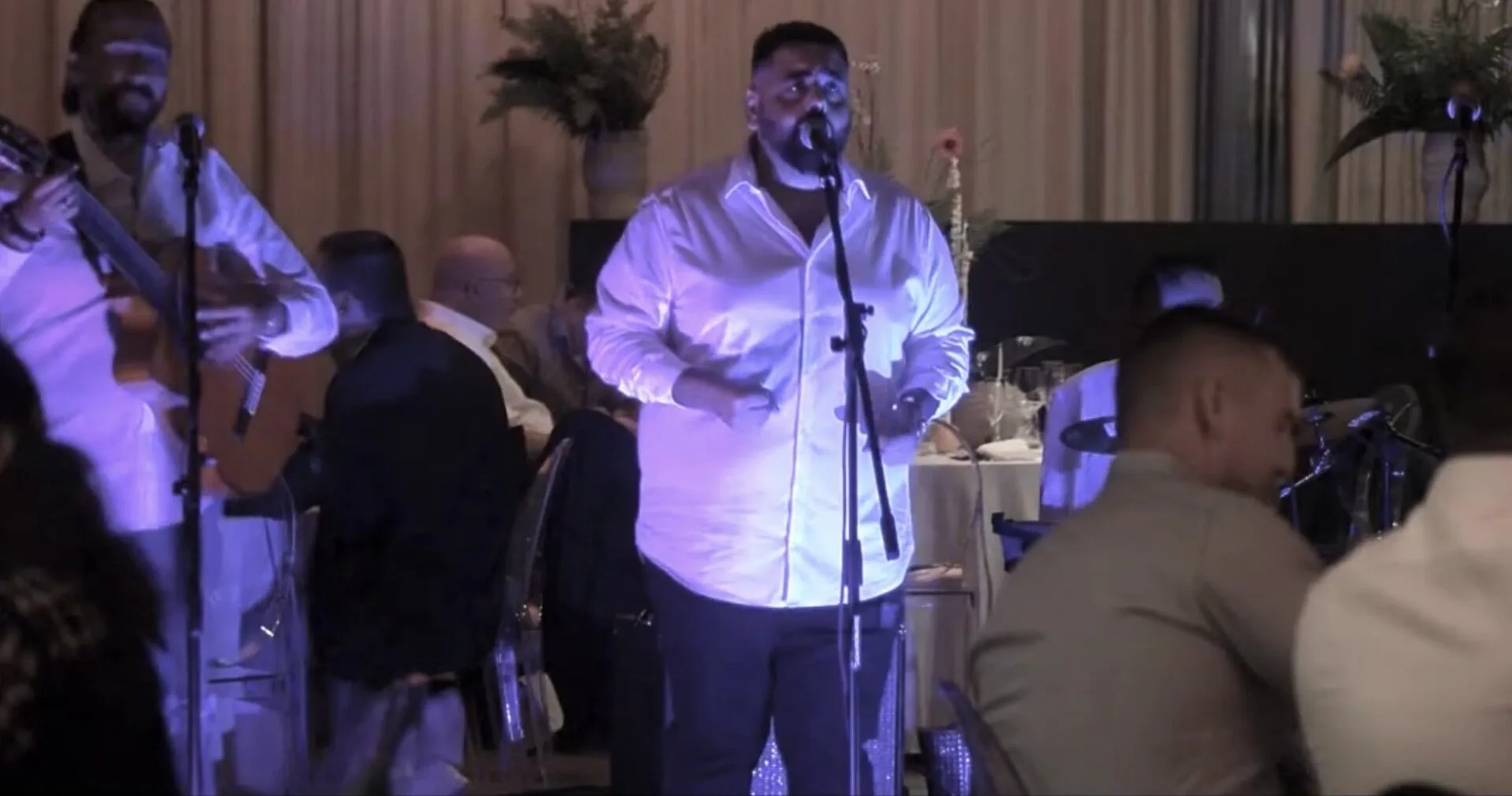 Un cantante con camisa blanca canta frente a un micrófono en un salón de celebración; a su izquierda se ve un guitarrista con camisa blanca y, al fondo, mesas con invitados sentados. La escena transmite una rumba que arranca sonrisas en tu boda en Tarragona, con ambiente de fiesta y música española.