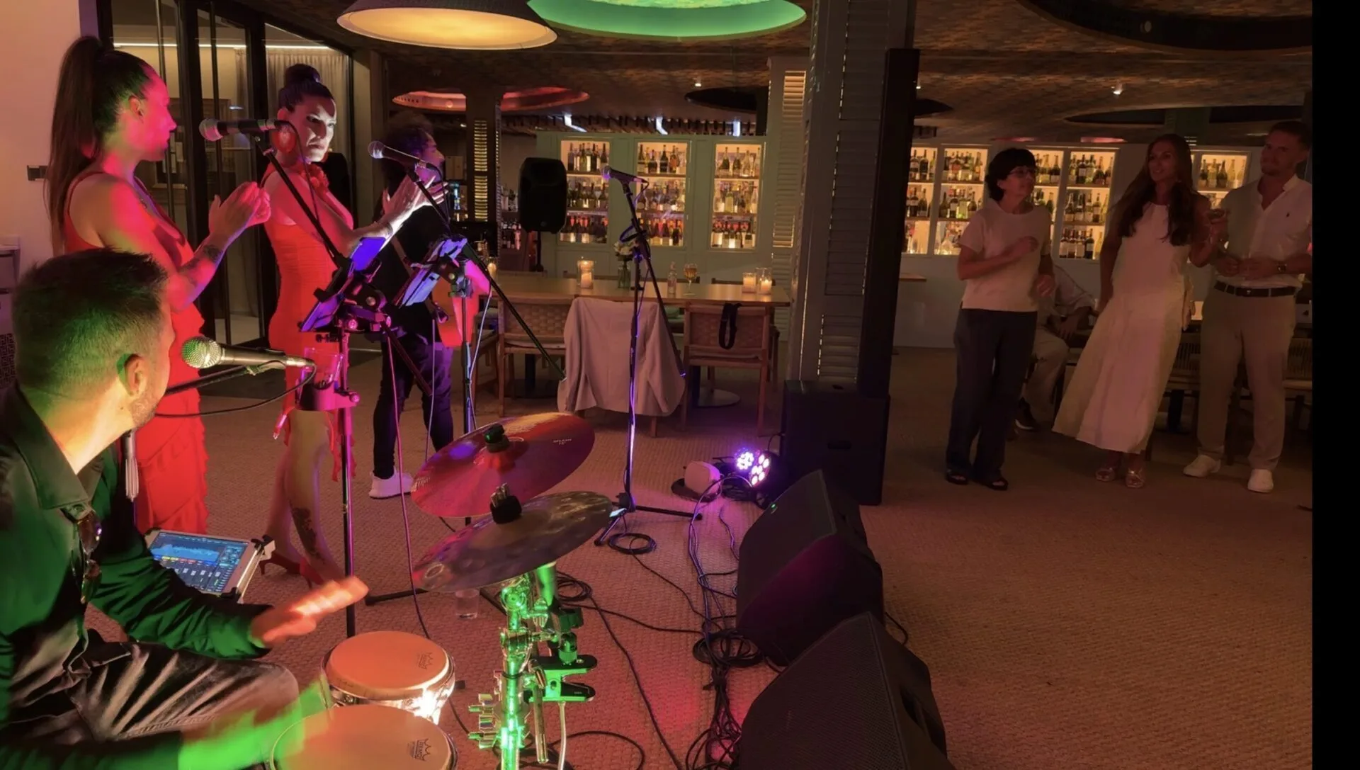 En el interior de un bar con estanterías de botellas al fondo, dos cantantes con micrófonos y vestidos rojos aplauden frente al público, mientras un percusionista toca en primer plano junto a una batería; a la derecha, varios asistentes —una mujer con vestido blanco y otras personas— bailan y observan. Escena adecuada para rumba fusión y pop flamenco para eventos de empresa en Barcelona.