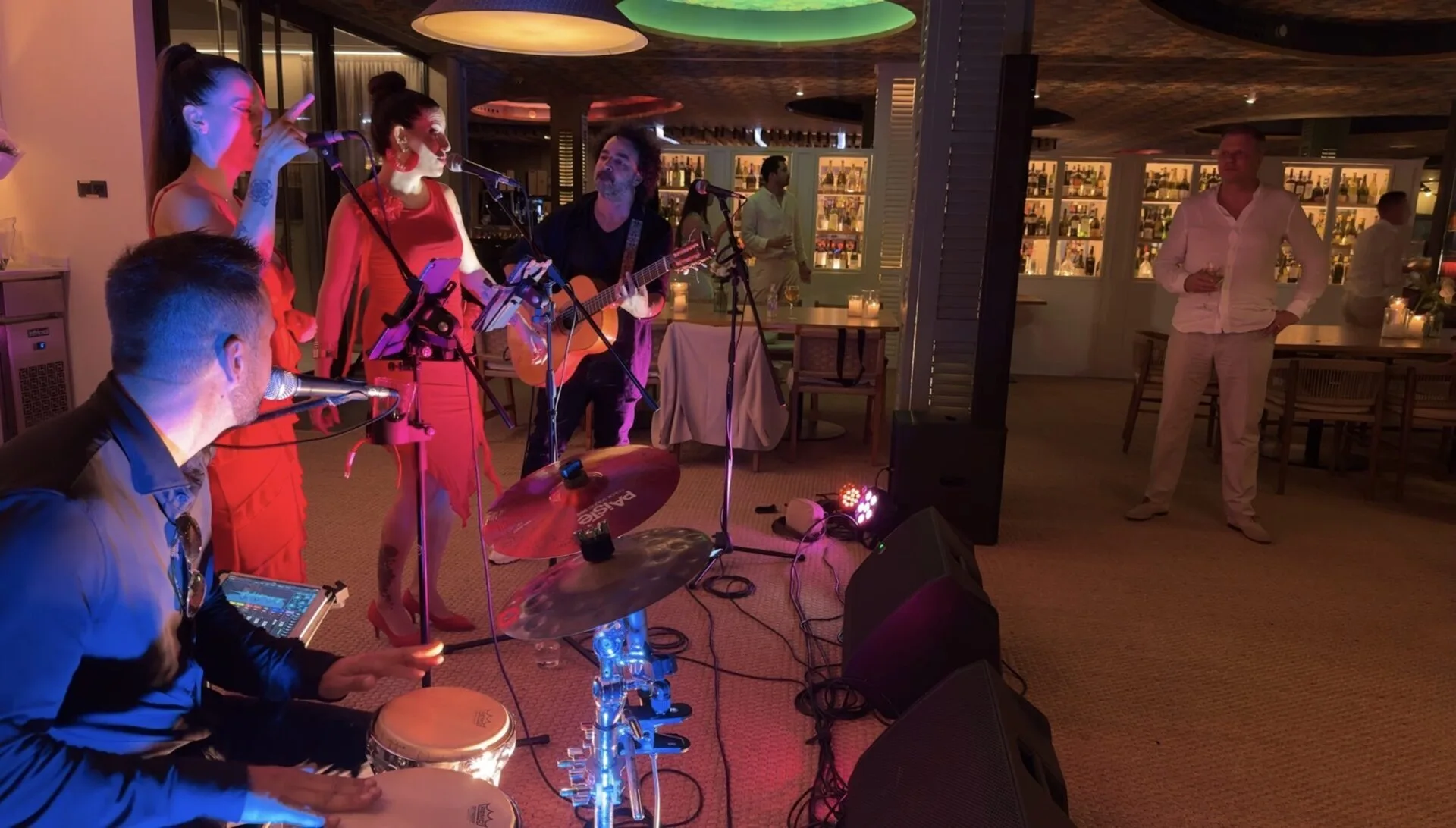 En un restaurante con luz cálida, dos cantantes con vestidos rojos cantan a dos micrófonos mientras un guitarrista toca sentado; en primer plano, un percusionista toca congas junto a un pequeño set de batería. Al fondo se ven botellas en estanterías y algunos asistentes mirando, en un ambiente de rumba fusión y pop flamenco para eventos de empresa en Barcelona.