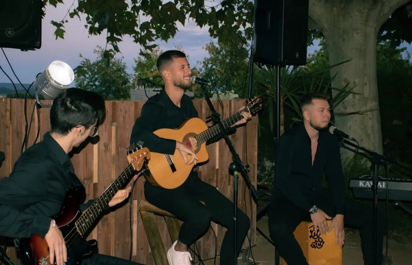 Trío de músicos con ropa negra tocando en un espacio exterior: el cantante en el centro toca una guitarra española y canta al micrófono, a la derecha un percusionista toca un cajón y a la izquierda un bajista acompaña; detrás hay una valla de madera, árboles y equipo de sonido, ambiente ideal para flamenquito y pop flamenco en Barcelona para cumpleaños con buen rollo.