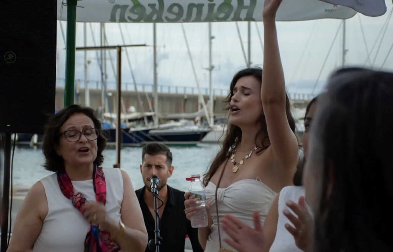 Varias personas cantan y aplauden en una terraza junto a un puerto con veleros; una mujer joven levanta el brazo y sostiene una botella de agua mientras, al fondo, un músico canta con un micrófono. Ambiente de celebración, ideal para flamenquito y pop flamenco en Barcelona para cumpleaños con buen rollo.