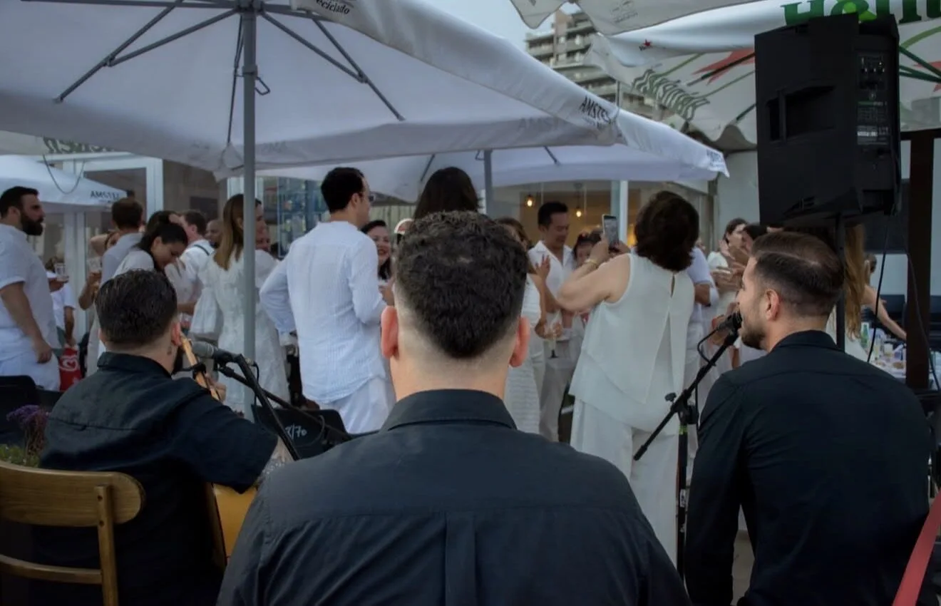 Vista desde detrás de tres músicos vestidos de negro con micrófonos y una guitarra, tocando ante un grupo de invitados en una terraza con sombrillas; la gente, vestida mayoritariamente de blanco, charla y baila en un ambiente festivo, ideal para flamenquito y pop flamenco en Barcelona para cumpleaños con buen rollo.