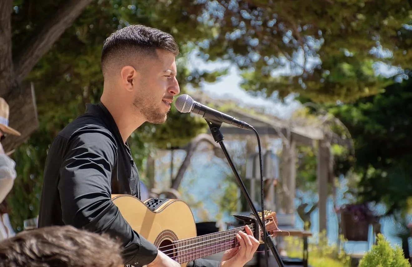 Primer plano de un cantante con camisa negra tocando una guitarra española y cantando en un micrófono, en una terraza exterior rodeada de árboles; una escena ideal para flamenquito y pop flamenco en Barcelona para cumpleaños con buen rollo.