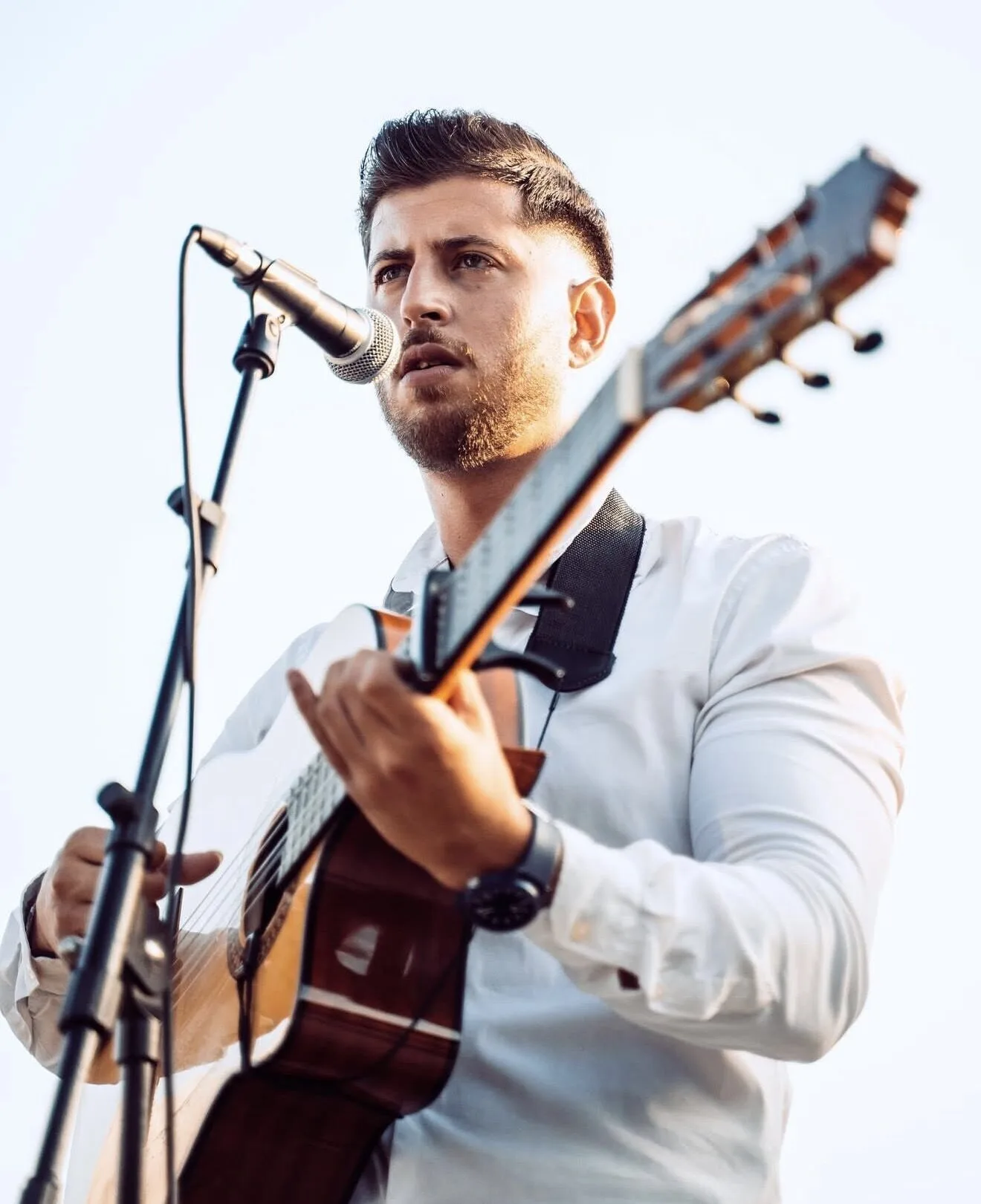 Primer plano de un cantante con barba y camisa blanca cantando ante un micrófono mientras sostiene y toca una guitarra española; la escena transmite un estilo de flamenquito y pop flamenco en Barcelona, ideal para cumpleaños con buen rollo.