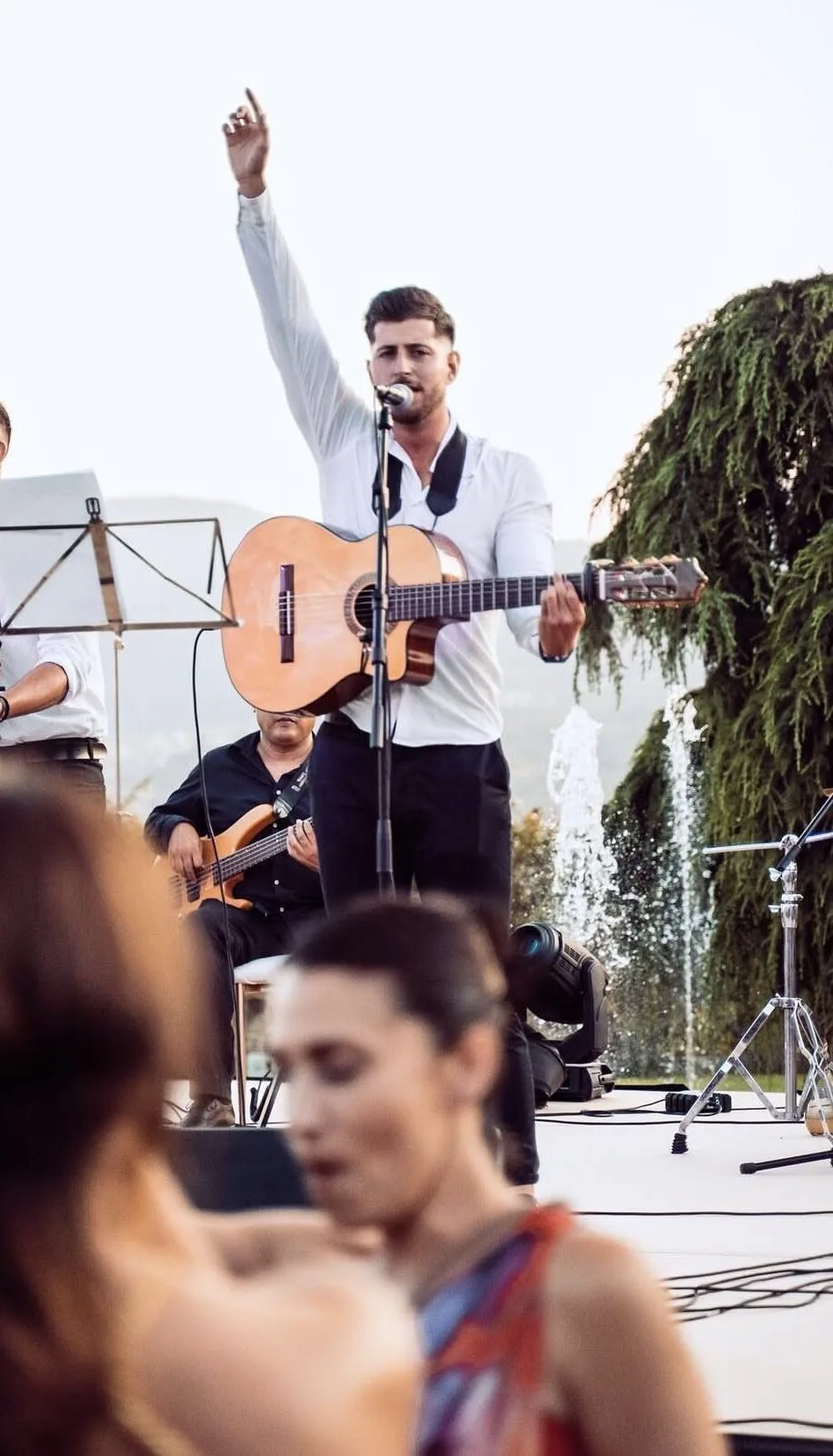 Cantante con camisa blanca tocando una guitarra española y cantando al micrófono con el brazo levantado, en un escenario al aire libre; detrás se ve un bajista sentado, una fuente de agua y árboles, con gente desenfocada en primer plano, ambiente de flamenquito y pop flamenco en Barcelona para cumpleaños con buen rollo.
