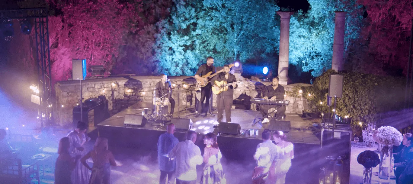 Grupo de rumba tocando en un escenario al aire libre de noche, con guitarrista al frente, batería, teclista y bajista detrás; delante, varios invitados bailan y charlan bajo luces moradas y azules, creando una rumba que arranca sonrisas en tu boda en Tarragona