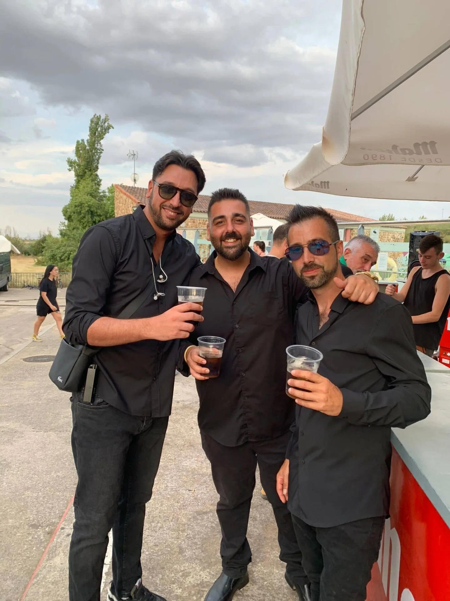 Tres hombres vestidos de negro posan sonrientes en una terraza al aire libre y levantan vasos de bebida, con gente y una sombrilla de bar al fondo; una escena de rumba que arranca sonrisas en tu boda en Tarragona, con un ambiente festivo y cercano.